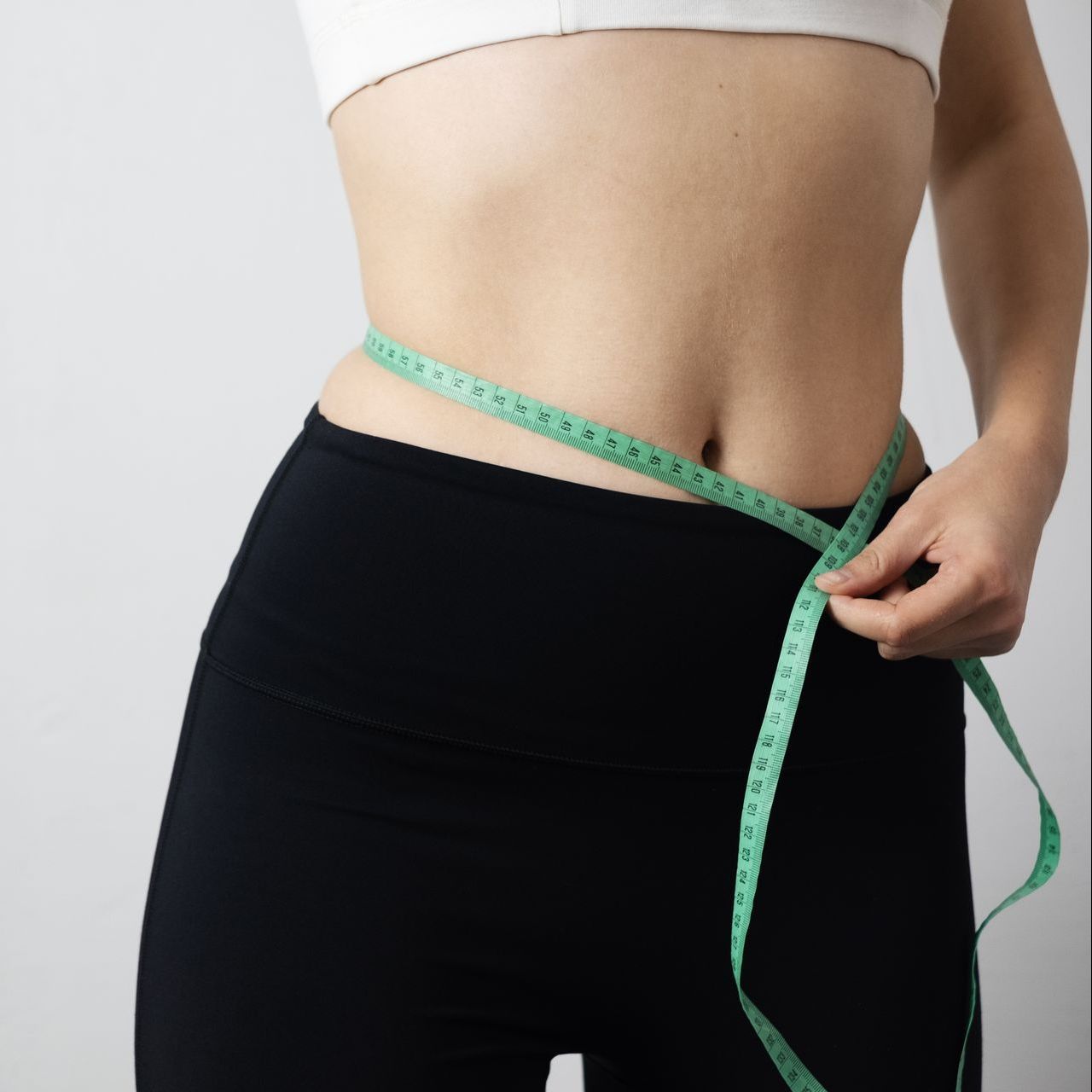Woman measuring waist with a green measuring tape, wearing a white top and black leggings.
