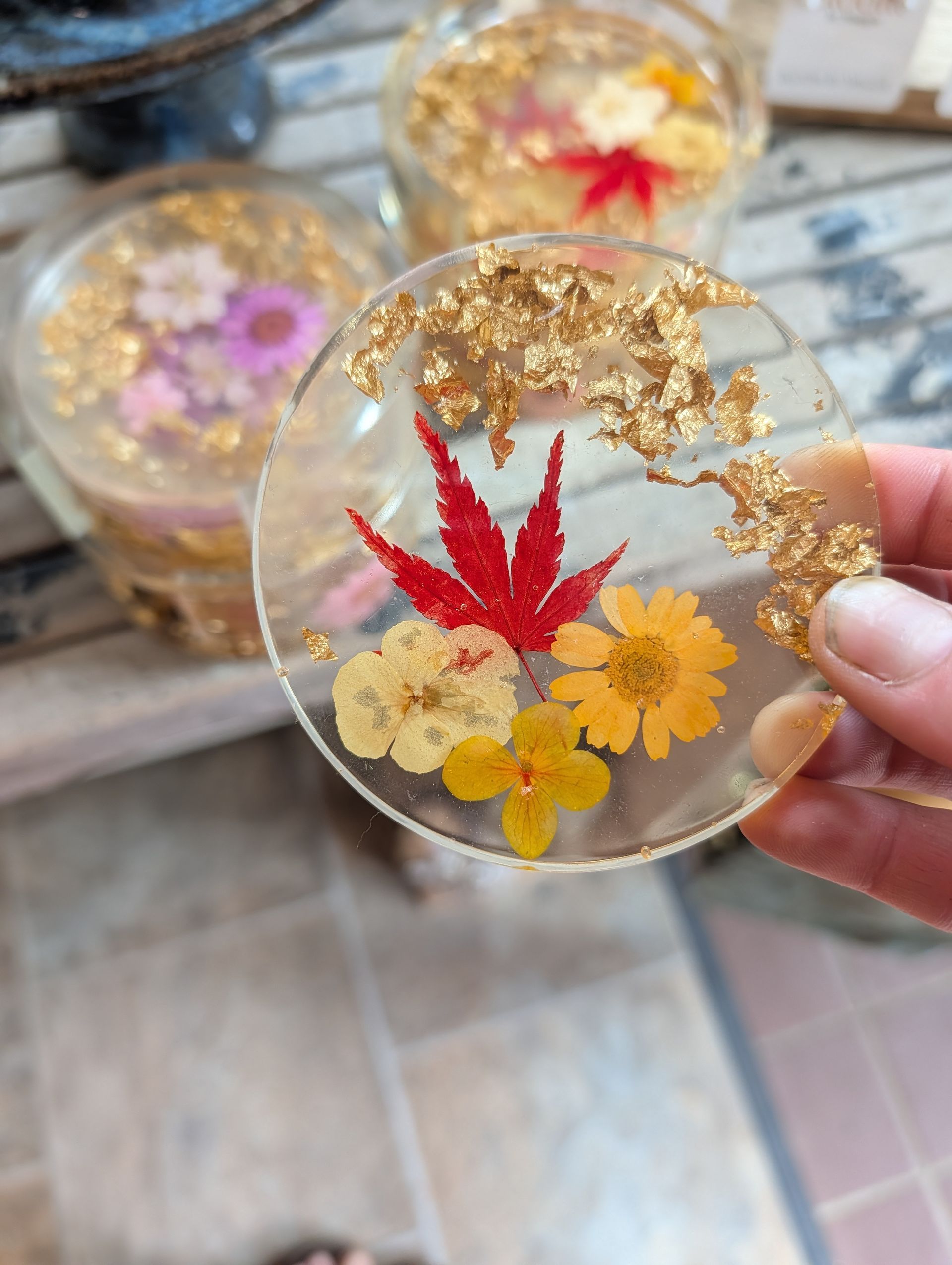 Hand holding a clear resin coaster with red and yellow pressed flowers and gold flakes
