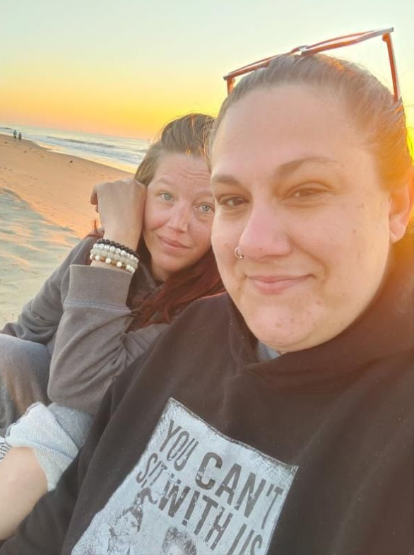 Two people selfie on a beach at sunset, with warm orange sky and ocean in the background.