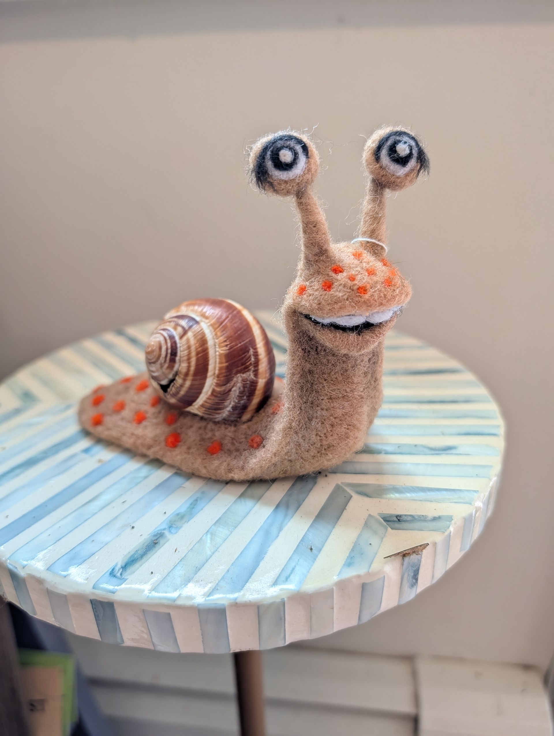 Cute handmade snail figurine with googly eyes, brown shell, and orange spots on a blue-and-white striped surface