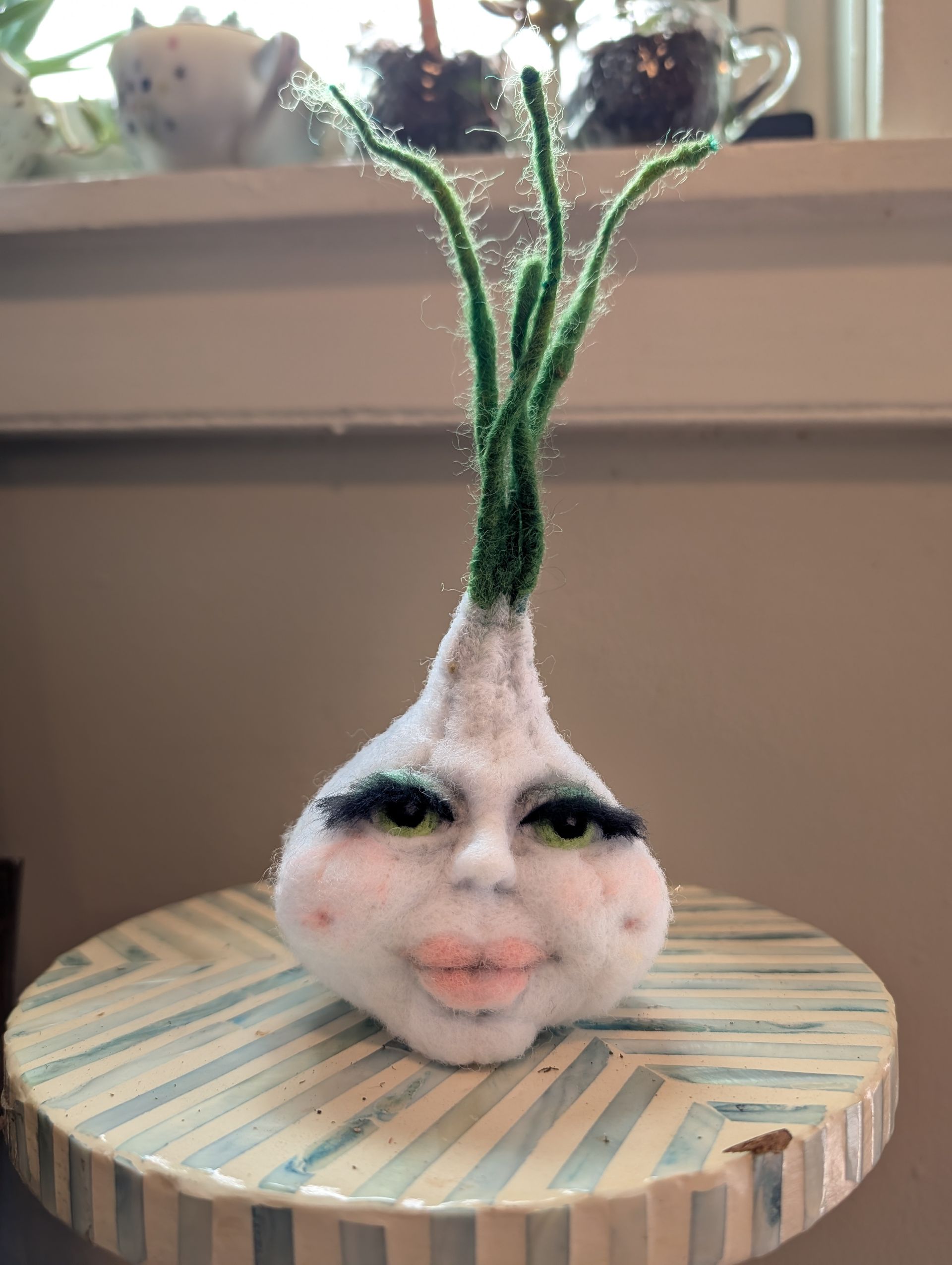 Small felt plant with human face and green stems on a striped round wooden base near a window.