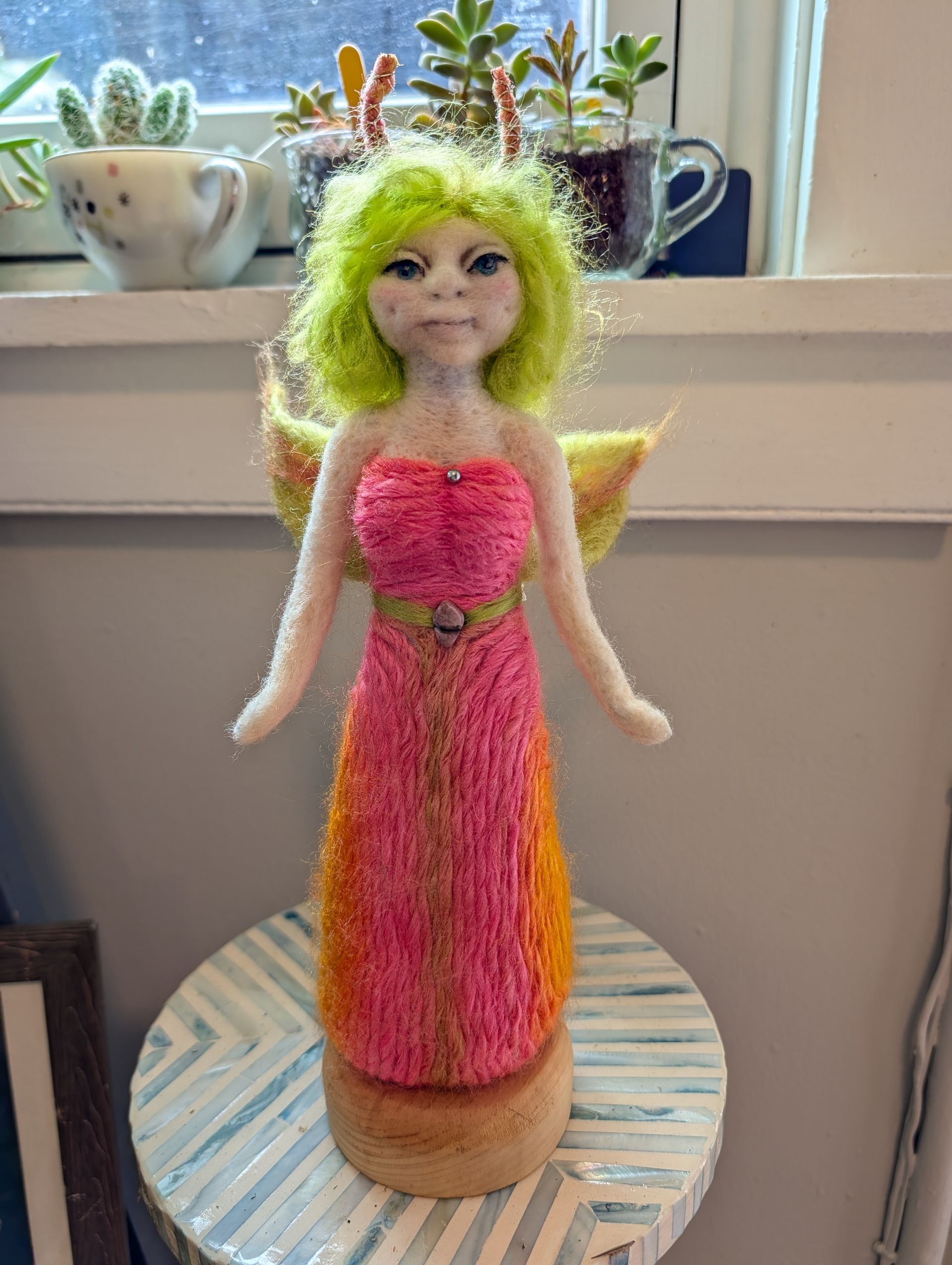 Pink-and-orange yarn doll with green hair on a striped round base by a windowsill, with potted plants behind