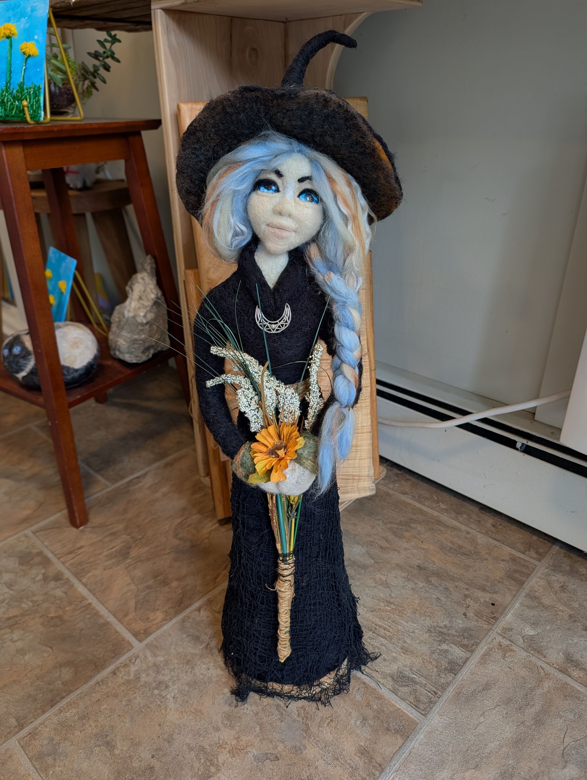 Black-clad doll with long pale braids, a hat, and a sunflower necklace standing on a tiled floor indoors