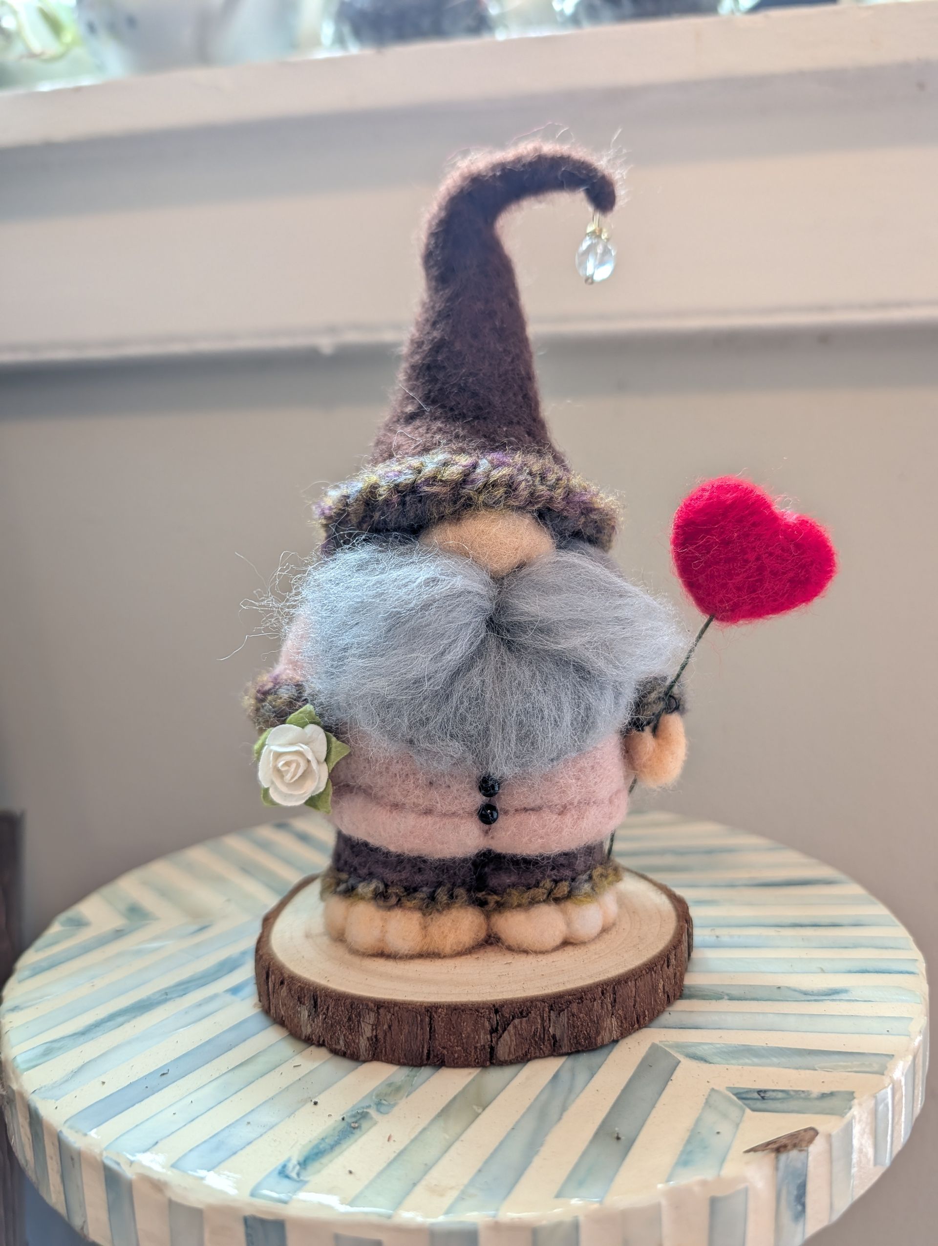 Gray felt gnome with tall brown hat, holding a red heart, on a striped round base by a window