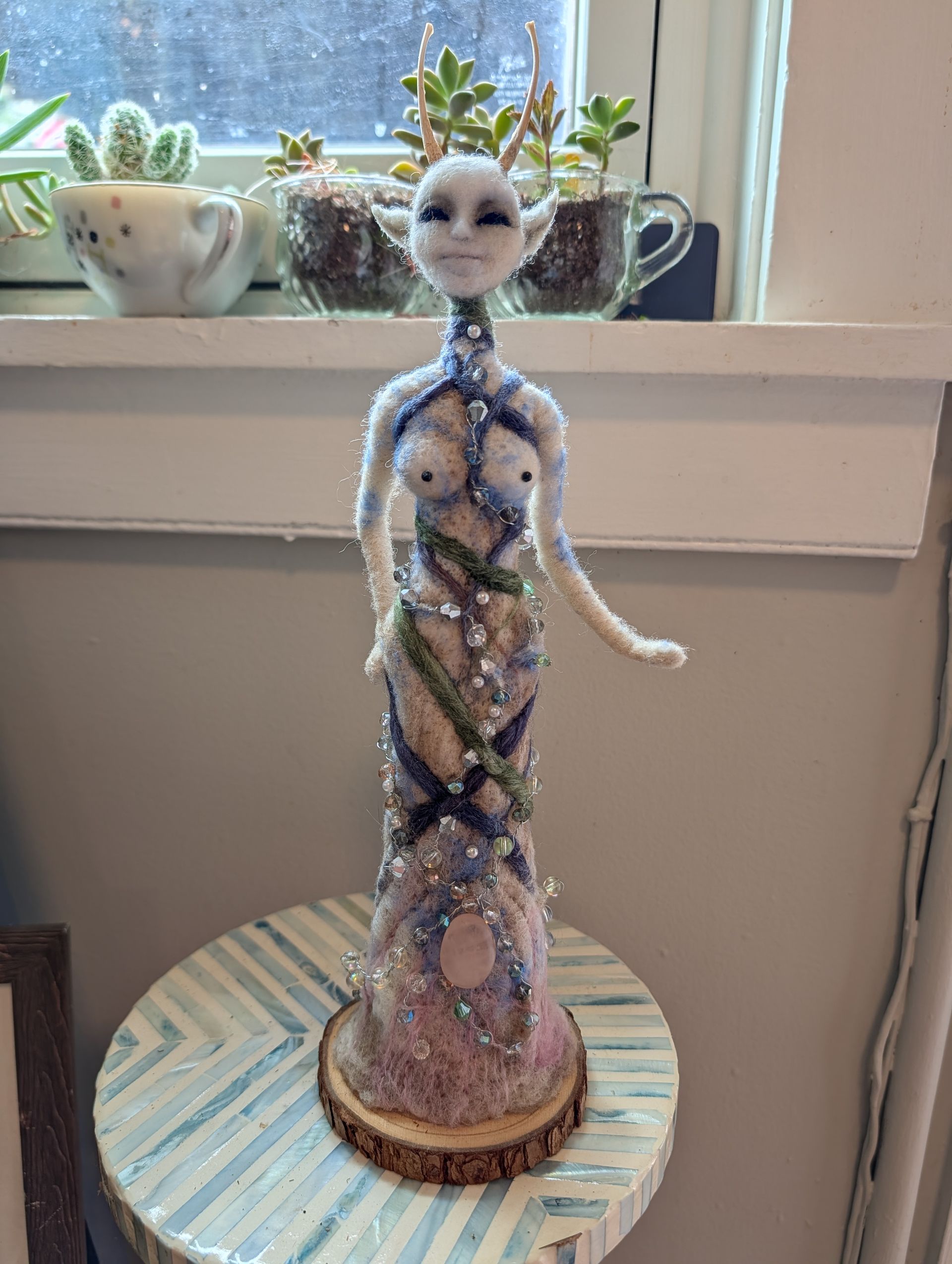 Decorative beaded figurine with a white face and blue gown on a striped table by a window.