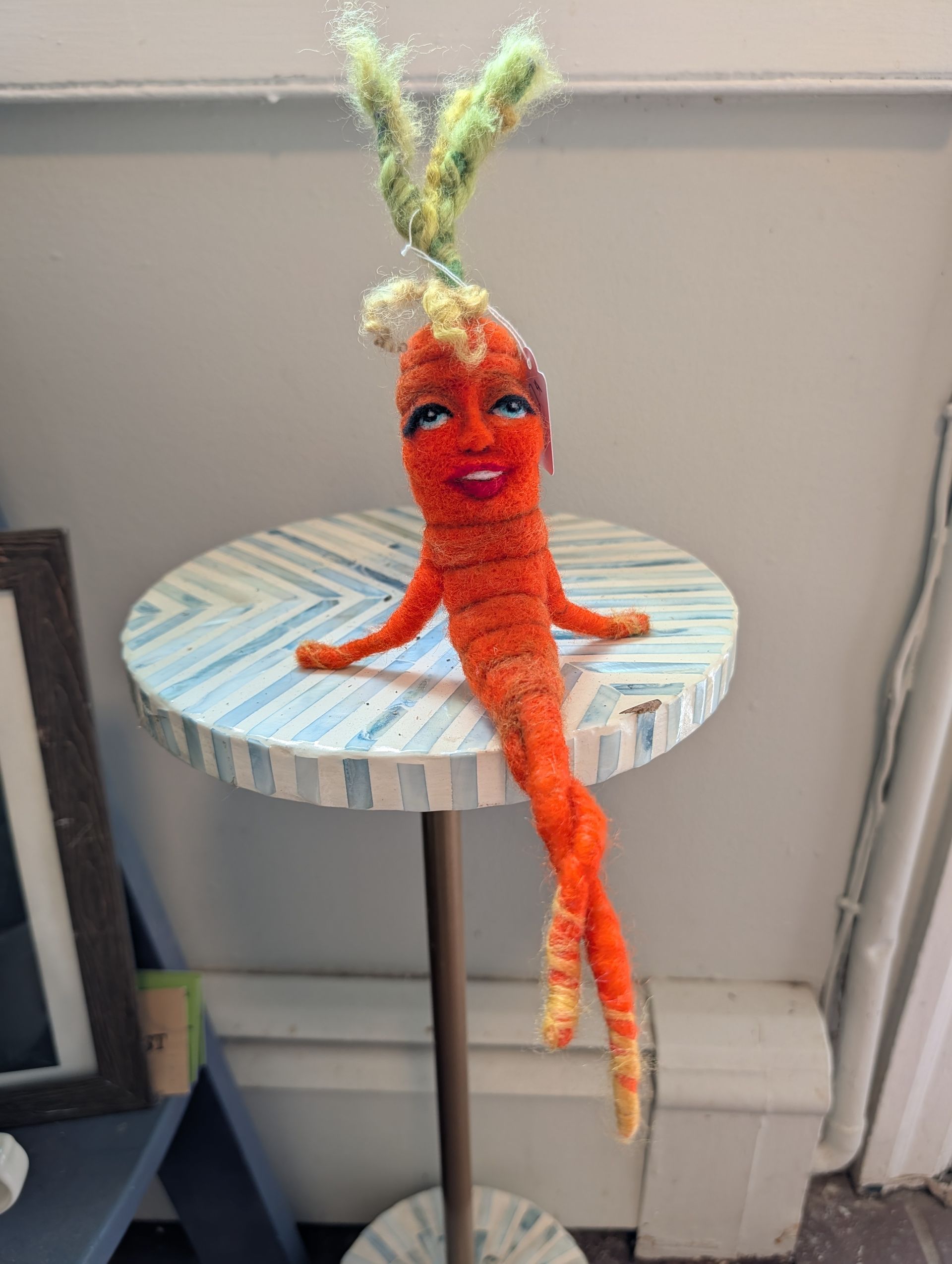 Orange crocheted figure with green yarn hair sitting on a round striped table by a wall