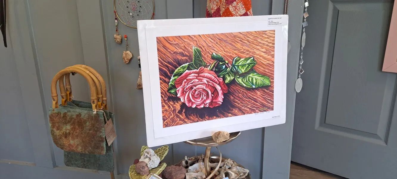 Painting of a pink rose on a wooden surface, displayed on a stand. An artisanal bag hangs to the left.