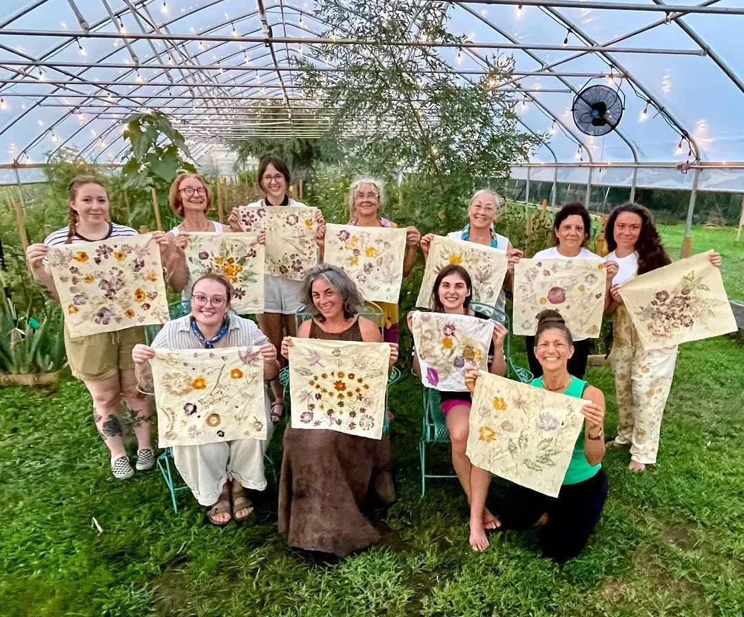 Group of people holding up decorated fabric squares in a greenhouse, smiling, lit by natural light.