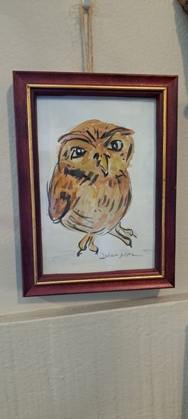 Owl painting in a dark red frame hanging on a wall by a string.