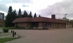 Single-story brown building with a sloped roof and brick chimney in a parking lot, trees behind it.