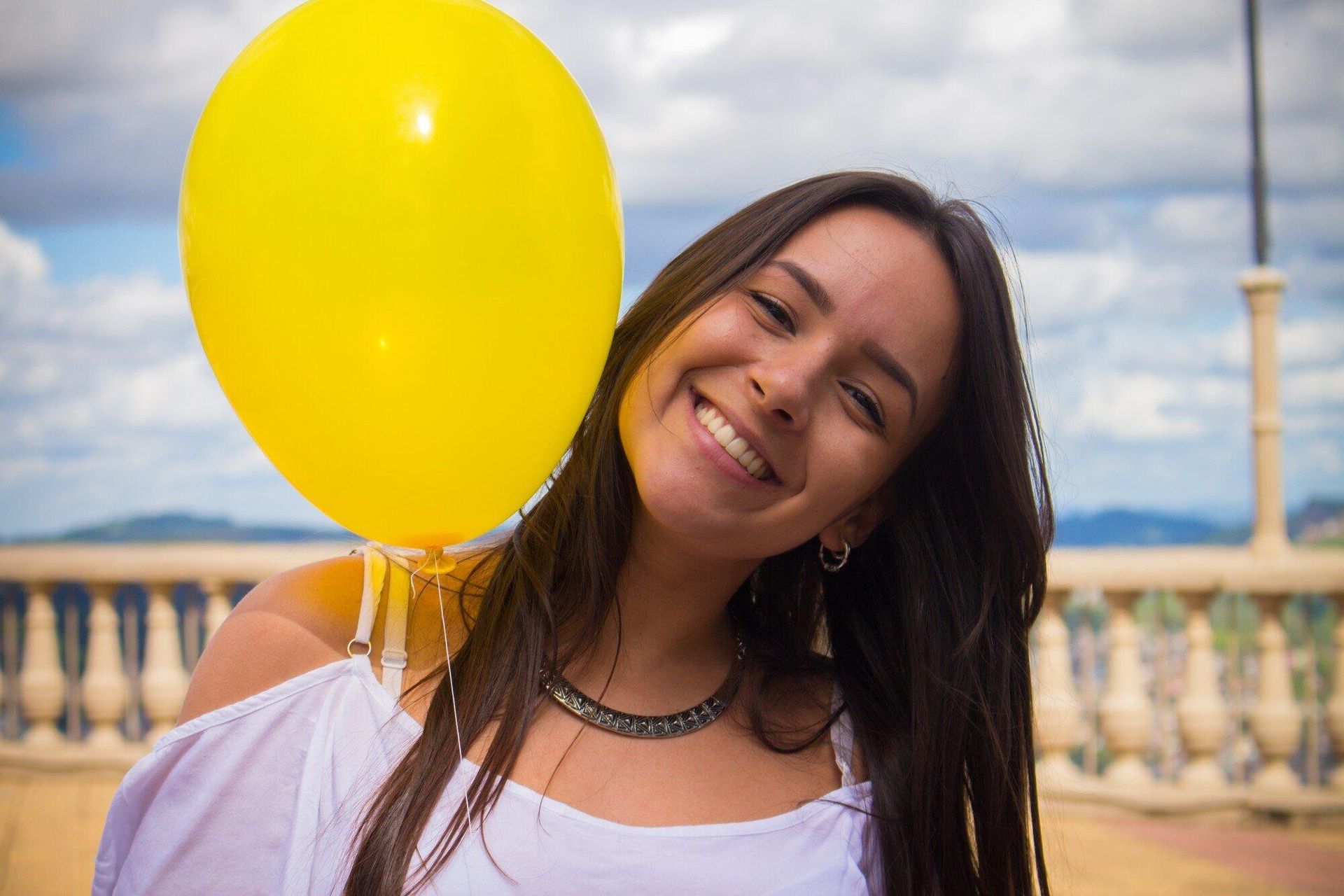 Smiling woman holding a yellow balloon outdoors by a seaside railing