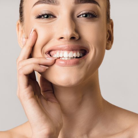 Woman smiling, touching cheek, showing white teeth, neutral background.