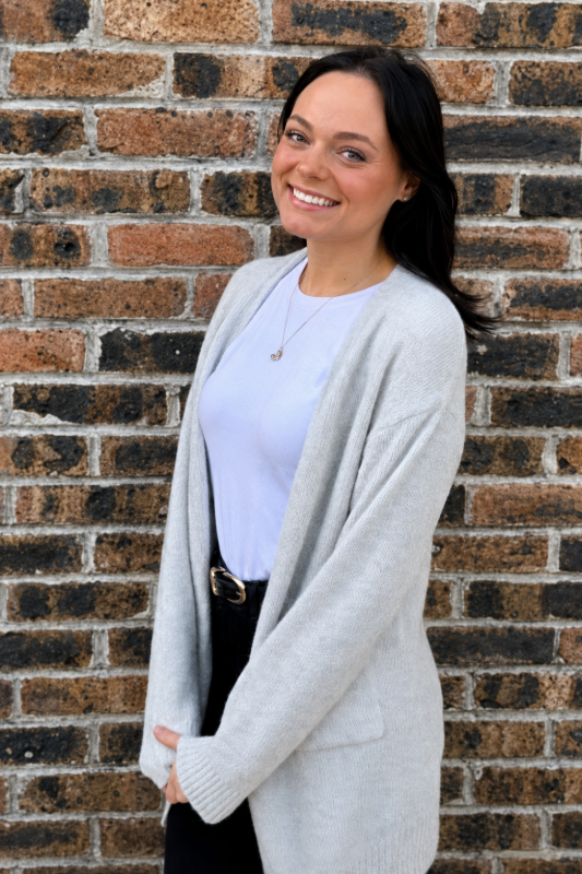 Smiling person in a light gray cardigan and white shirt standing against a brick wall