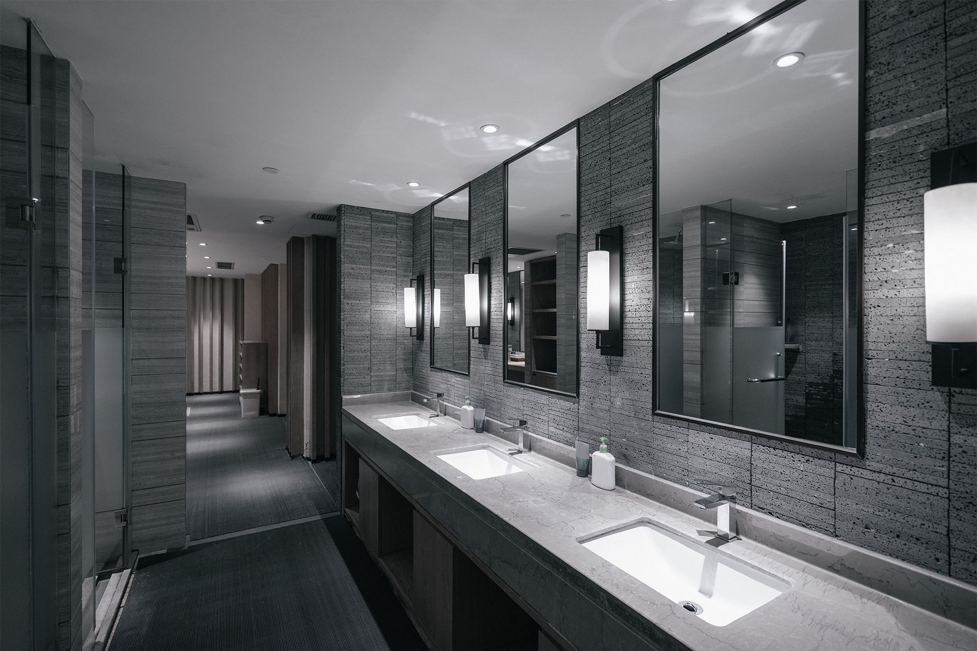 Modern public restroom with long granite sinks, mirrors, and gray tiled walls along a corridor.
