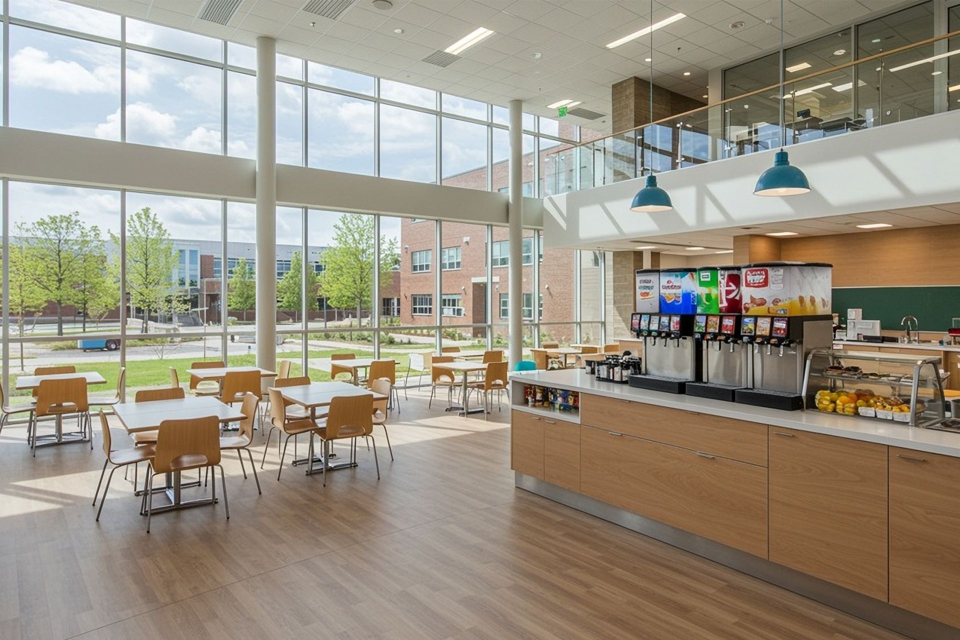 Bright cafeteria with large windows, wooden tables, and a coffee counter in a modern open dining space.