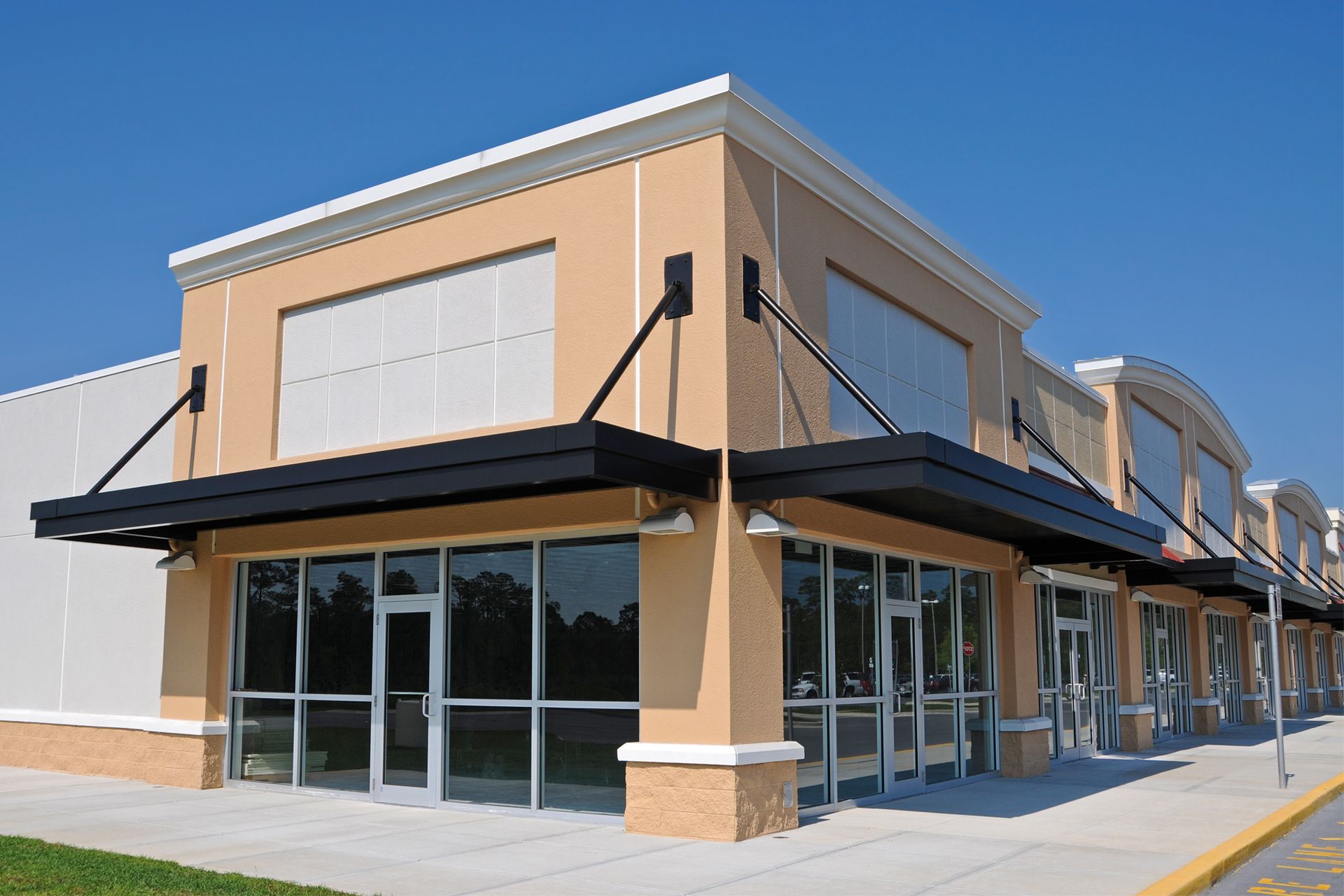 Empty storefronts in a tan shopping plaza with black awnings and glass windows under a clear sky.