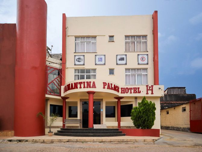 Vista frontal de um edifício de hotel em tons creme e vermelho com uma placa onde se lê "MANTINA PALM HOTEL".