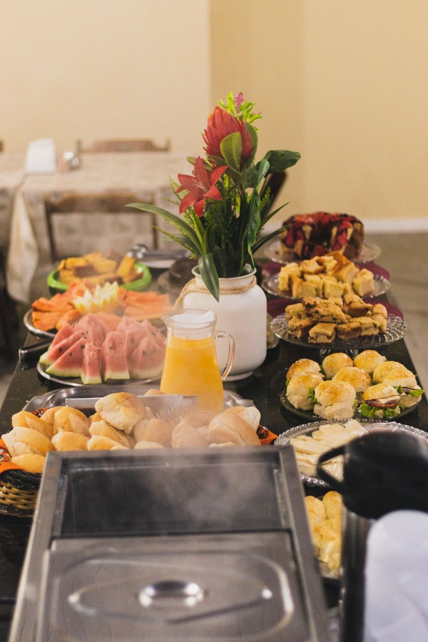 Mesa de buffet com travessas de frutas, doces e um arranjo de flores no centro da mesa, em uma sala de jantar.