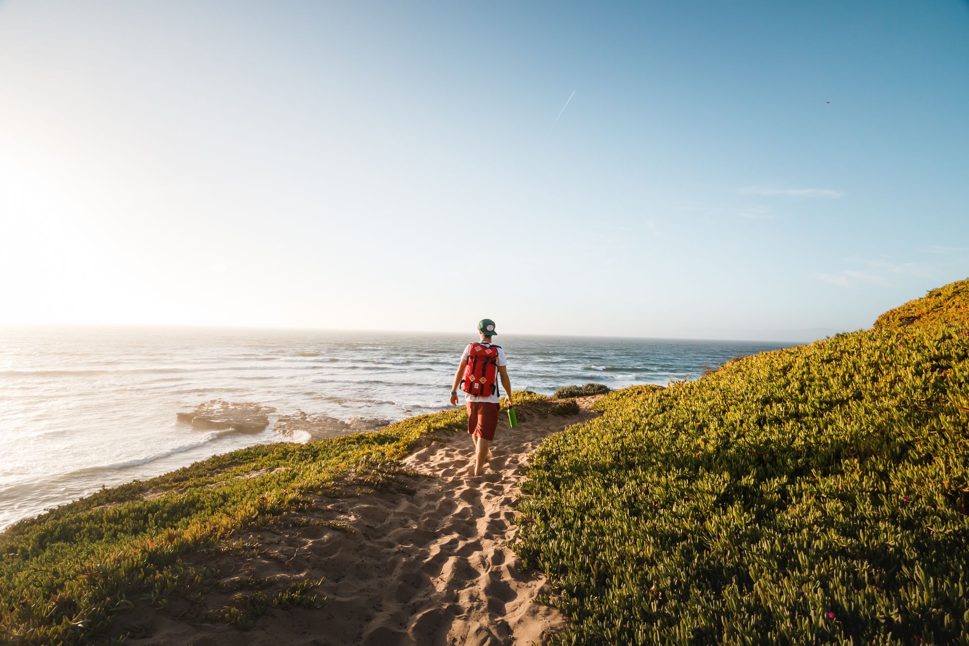 Hiker walking a coastal trail beside the ocean at sunset, with grassy cliffs and bright sky.