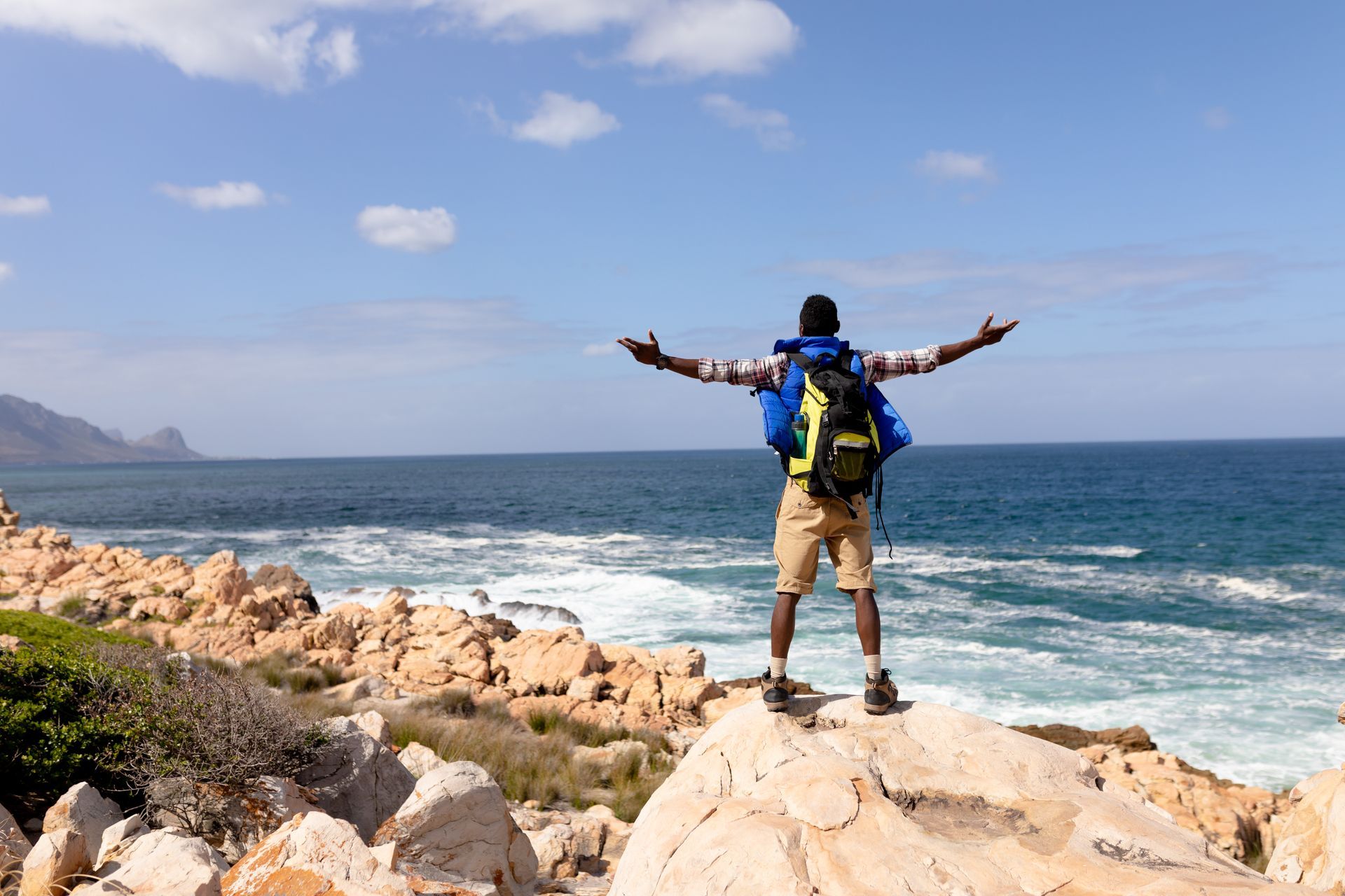 Backpacker stands on seaside rocks with arms outstretched, facing ocean waves under a blue sky.
