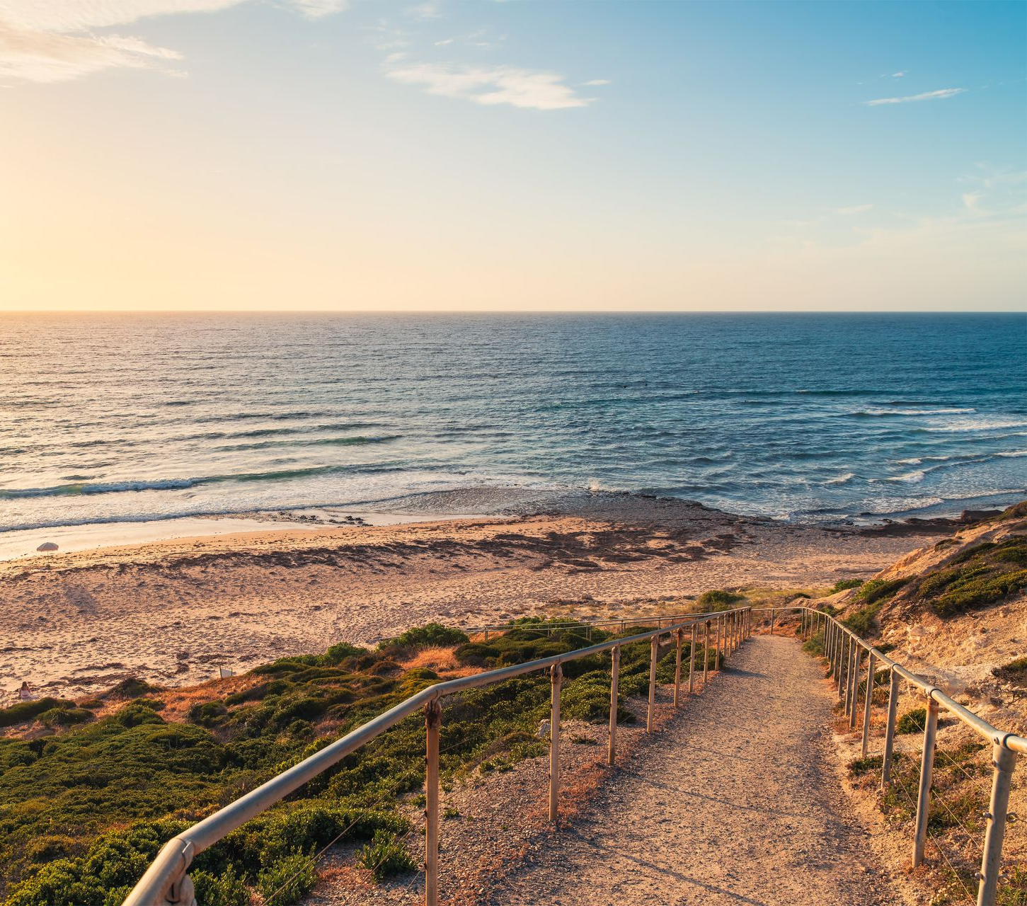 Sunlit coastal path leading to a rocky beach and calm blue ocean at sunset