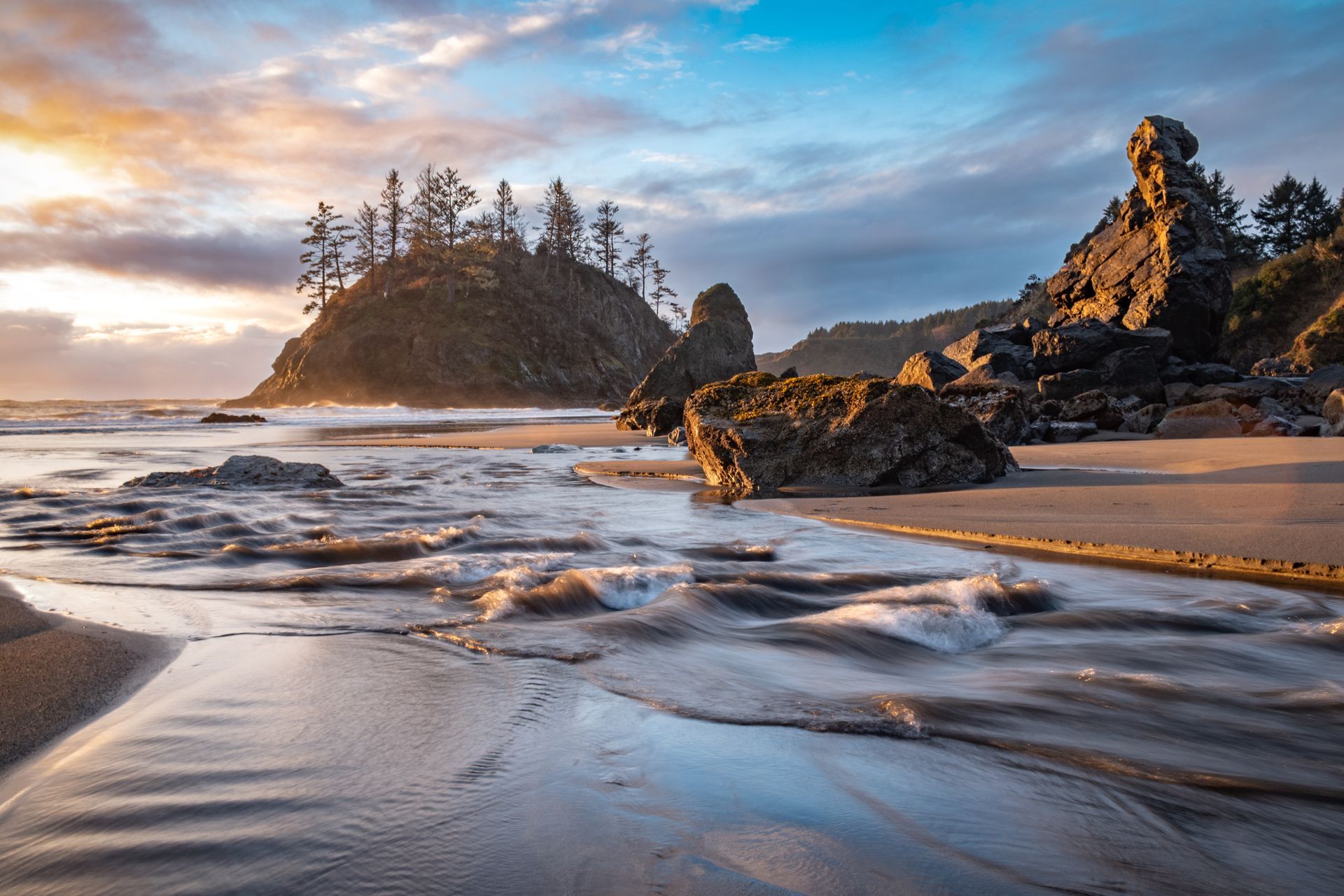 Sandy beach with waves, rocky outcrop, and pine-covered island at sunset