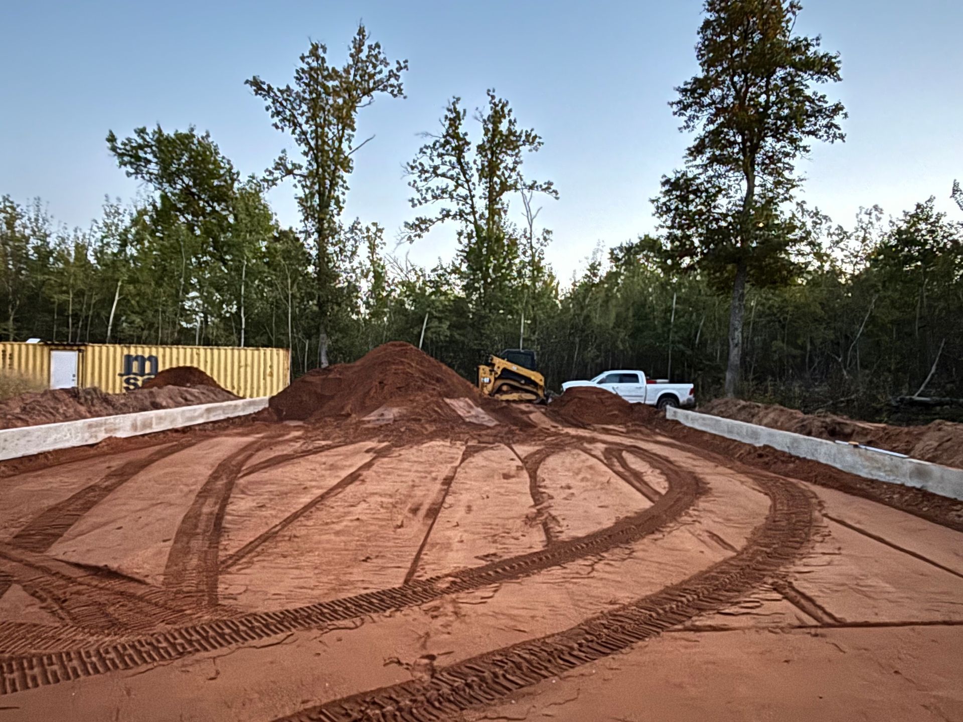 A construction site with reddish dirt, tire tracks, a bulldozer, a white pickup truck, and a yellow shipping container.