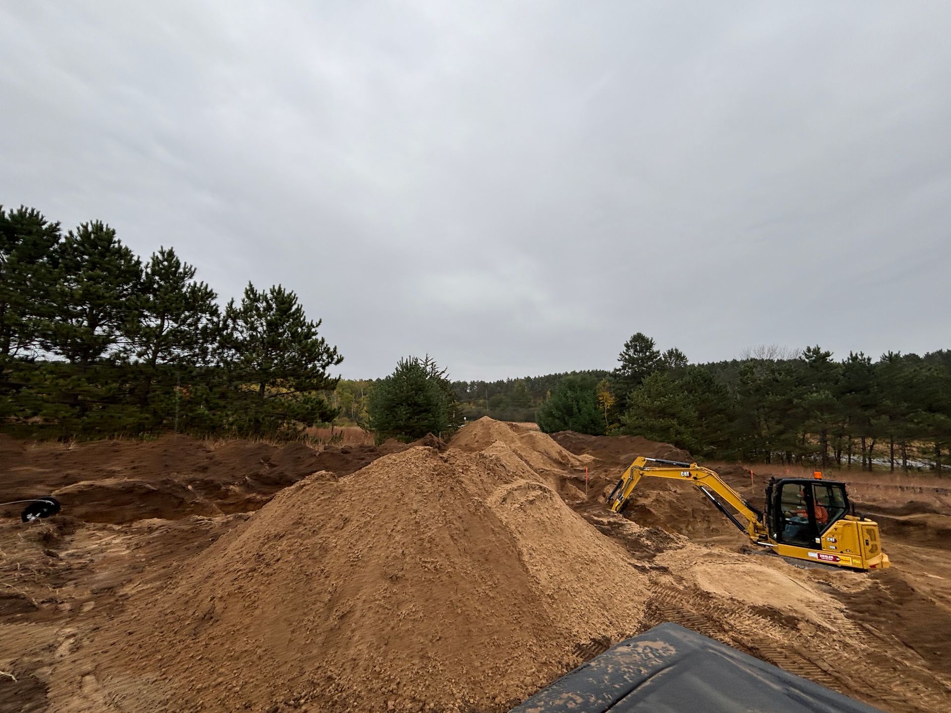 A yellow excavator works in a dirt-filled, excavated landscape under an overcast sky, with trees in the background.