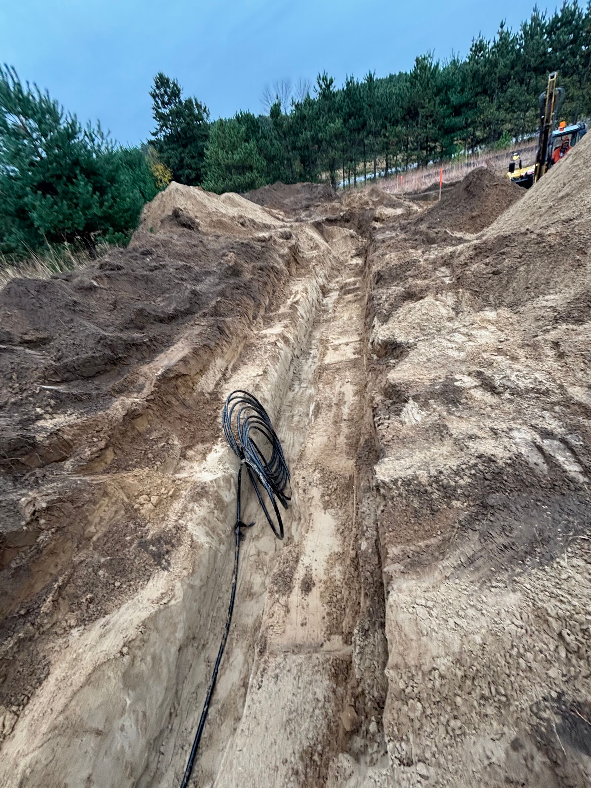 A trench is dug into sandy, brown soil, with a black utility cable running through it and a yellow excavator in the back.