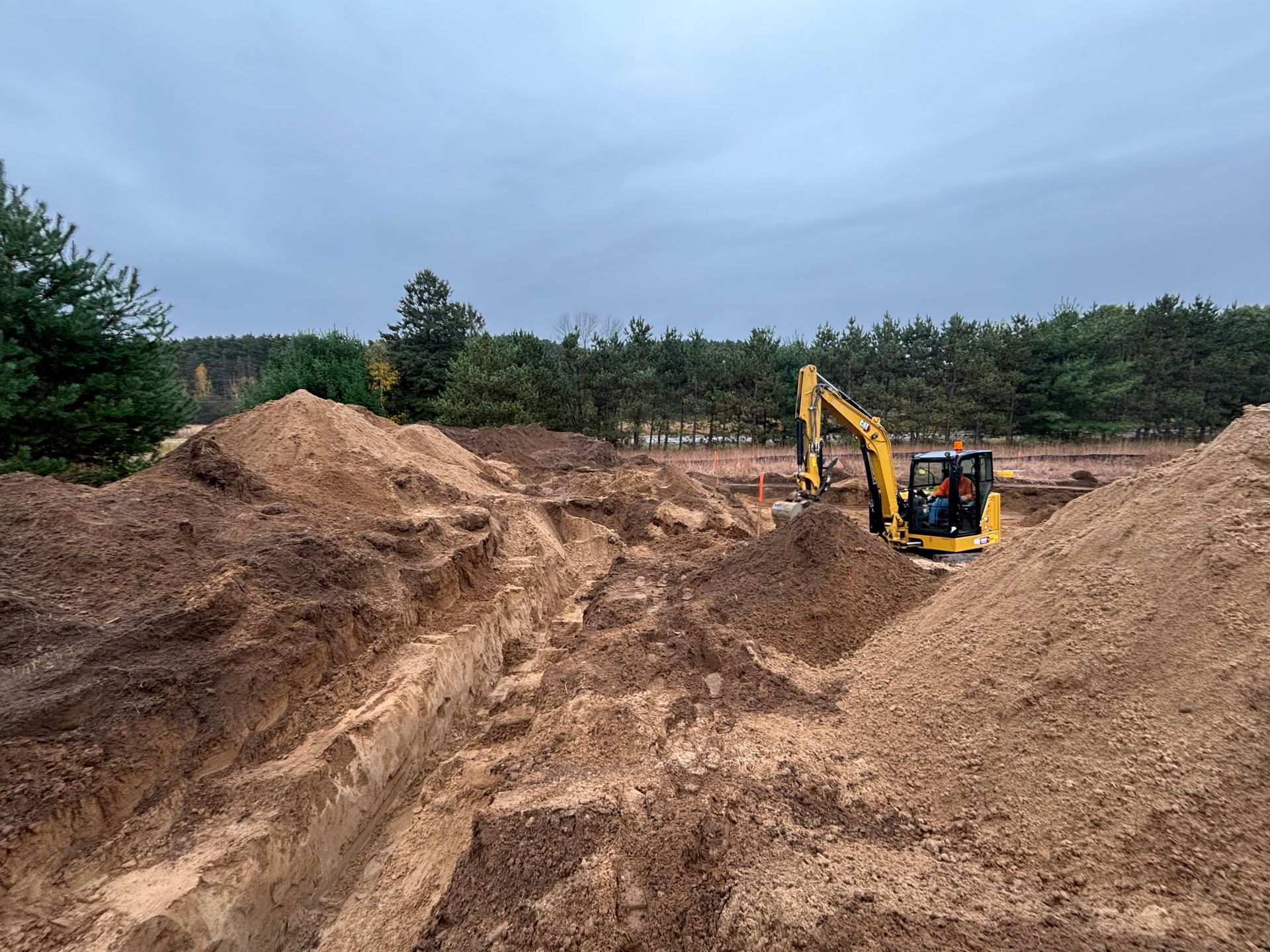 A yellow excavator sits in a dirt-filled construction site, moving soil near a long, deep trench under a cloudy sky.
