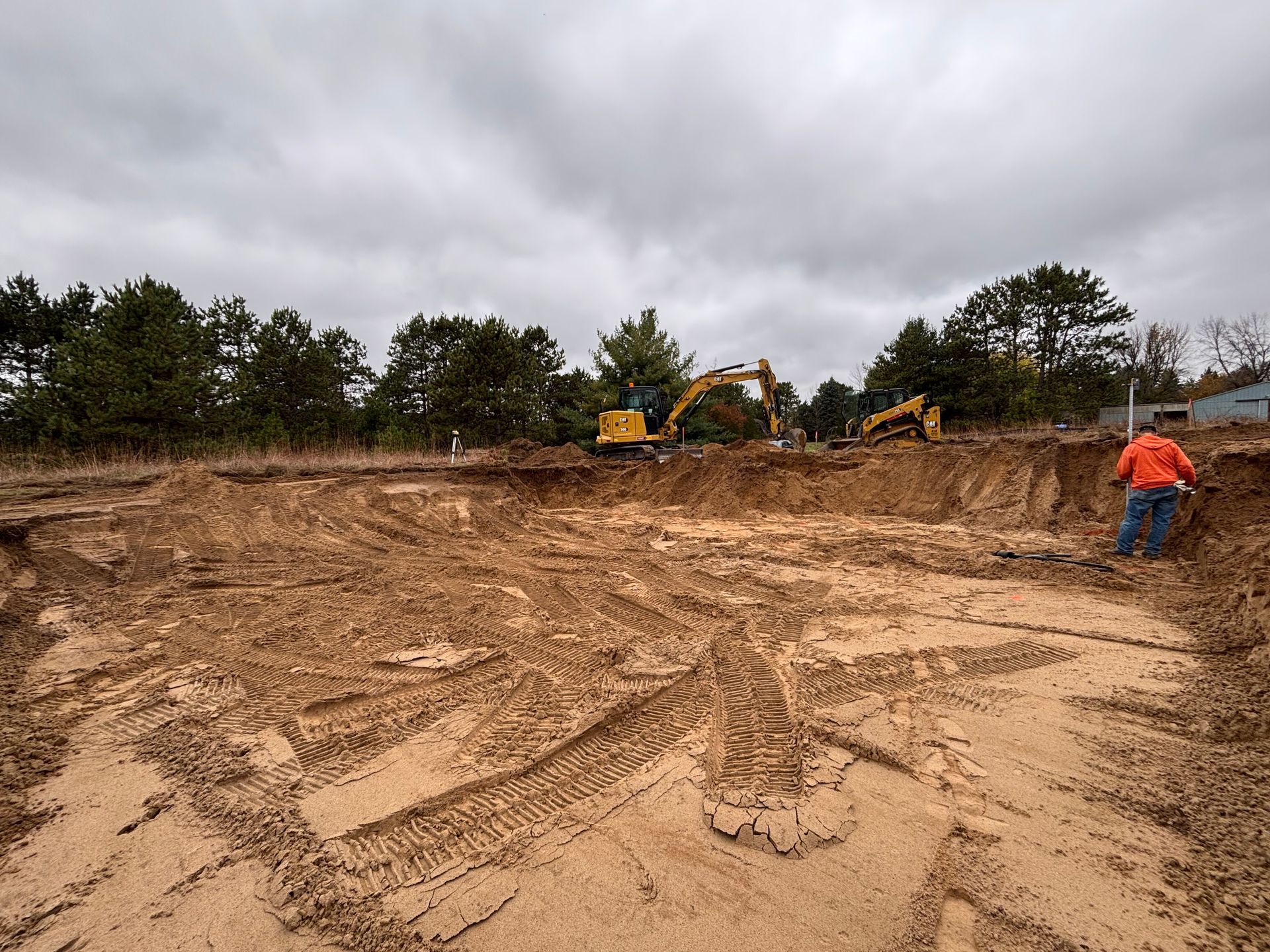 A construction site with a large dirt pit, heavy machinery, and a worker in an orange jacket on a cloudy day.