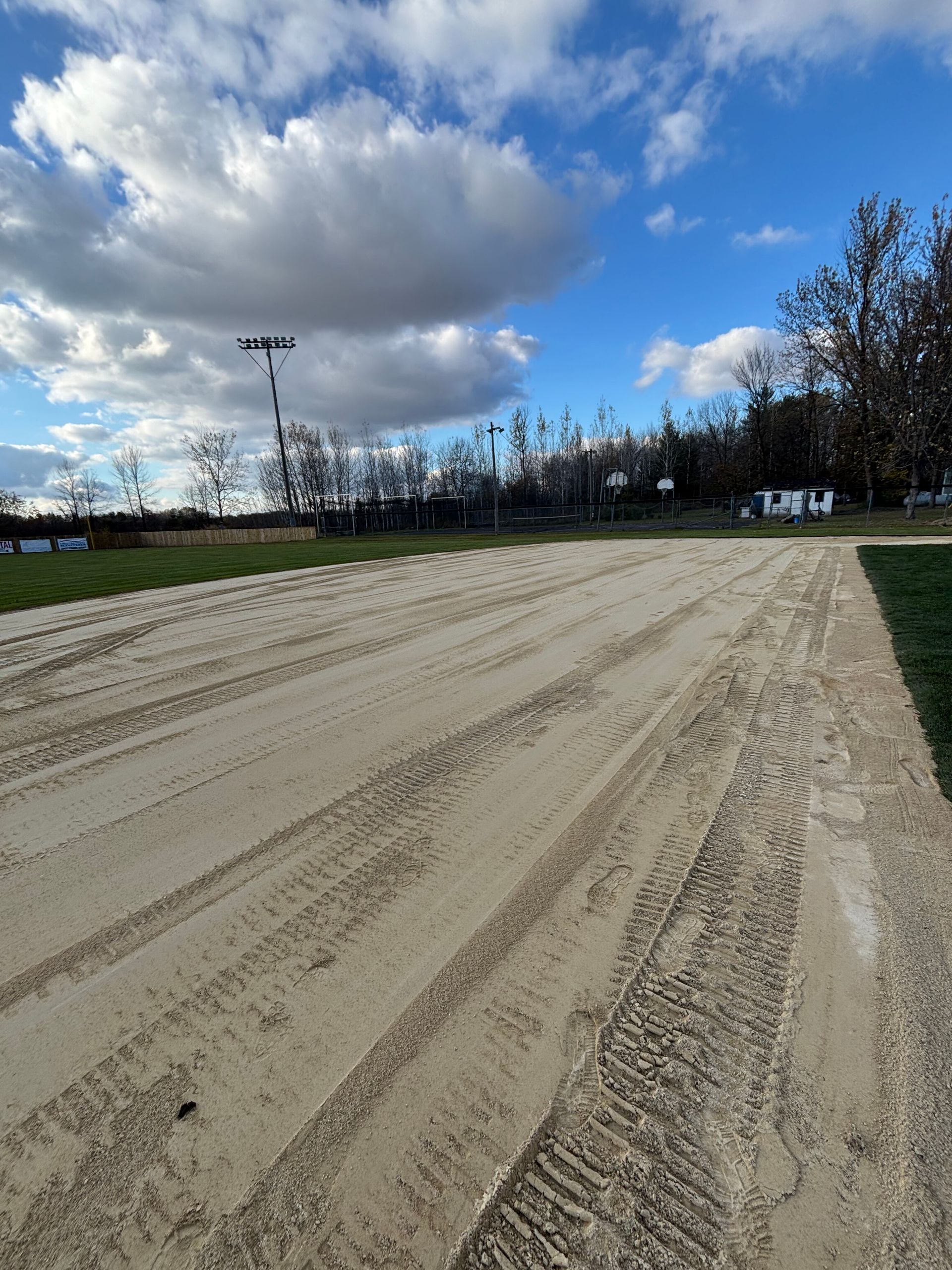 A wide, light-colored sand track with tire tracks, framed by green grass under a blue sky with scattered clouds.