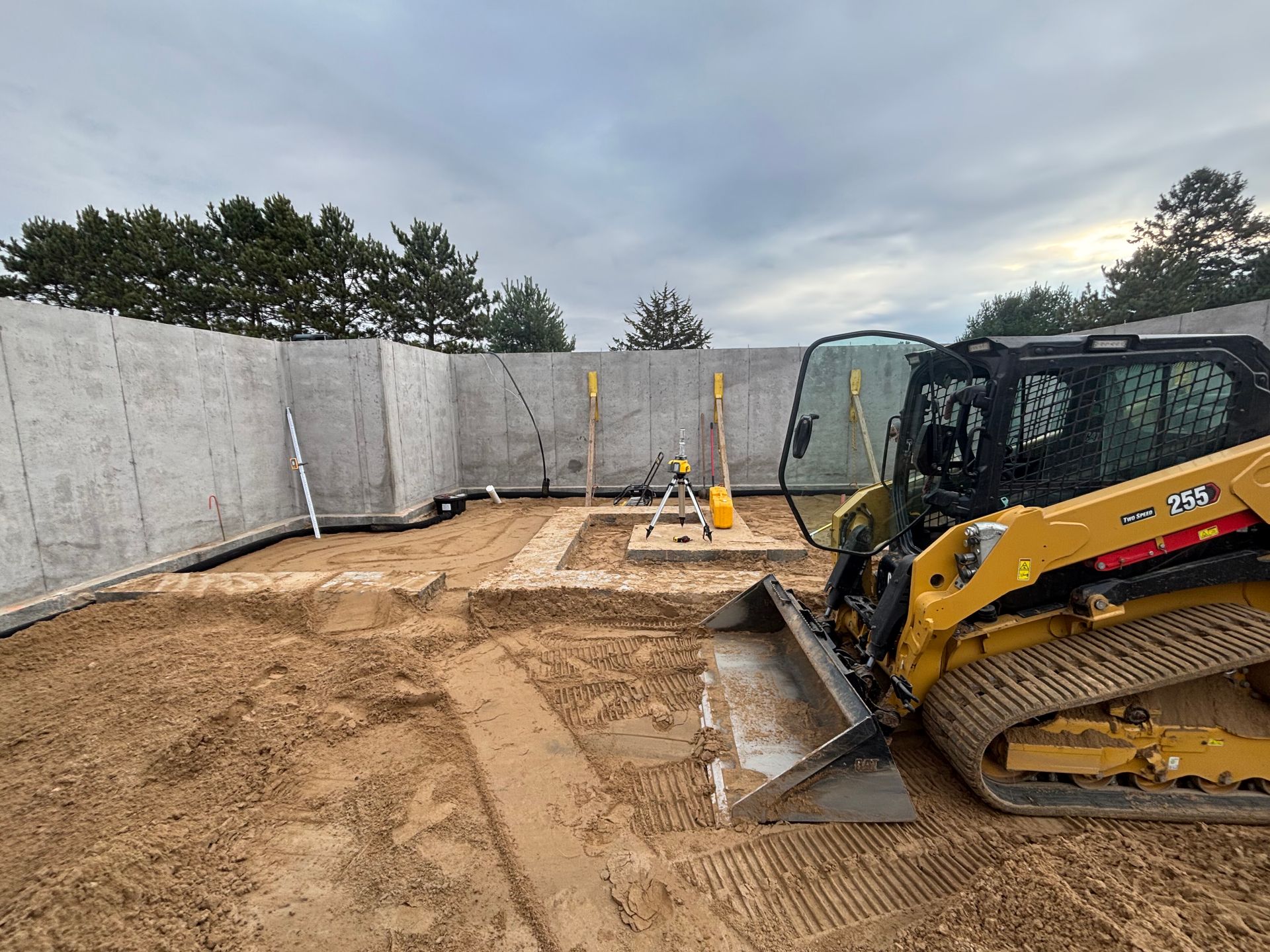 A yellow tracked skid steer parked on a sandy construction site with concrete foundation walls in the background.