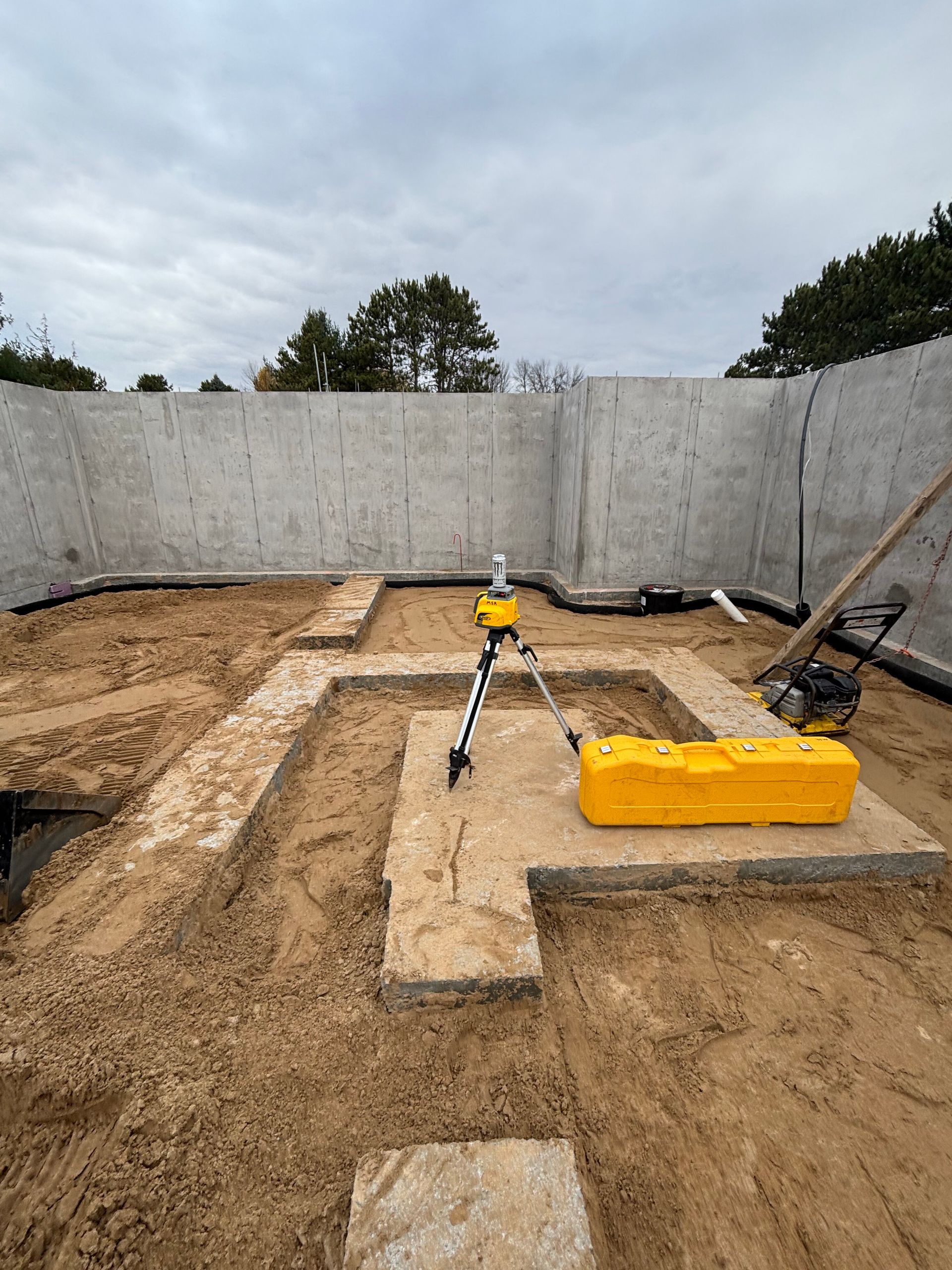 A laser level on a tripod sits atop a concrete footing inside a building foundation under construction.