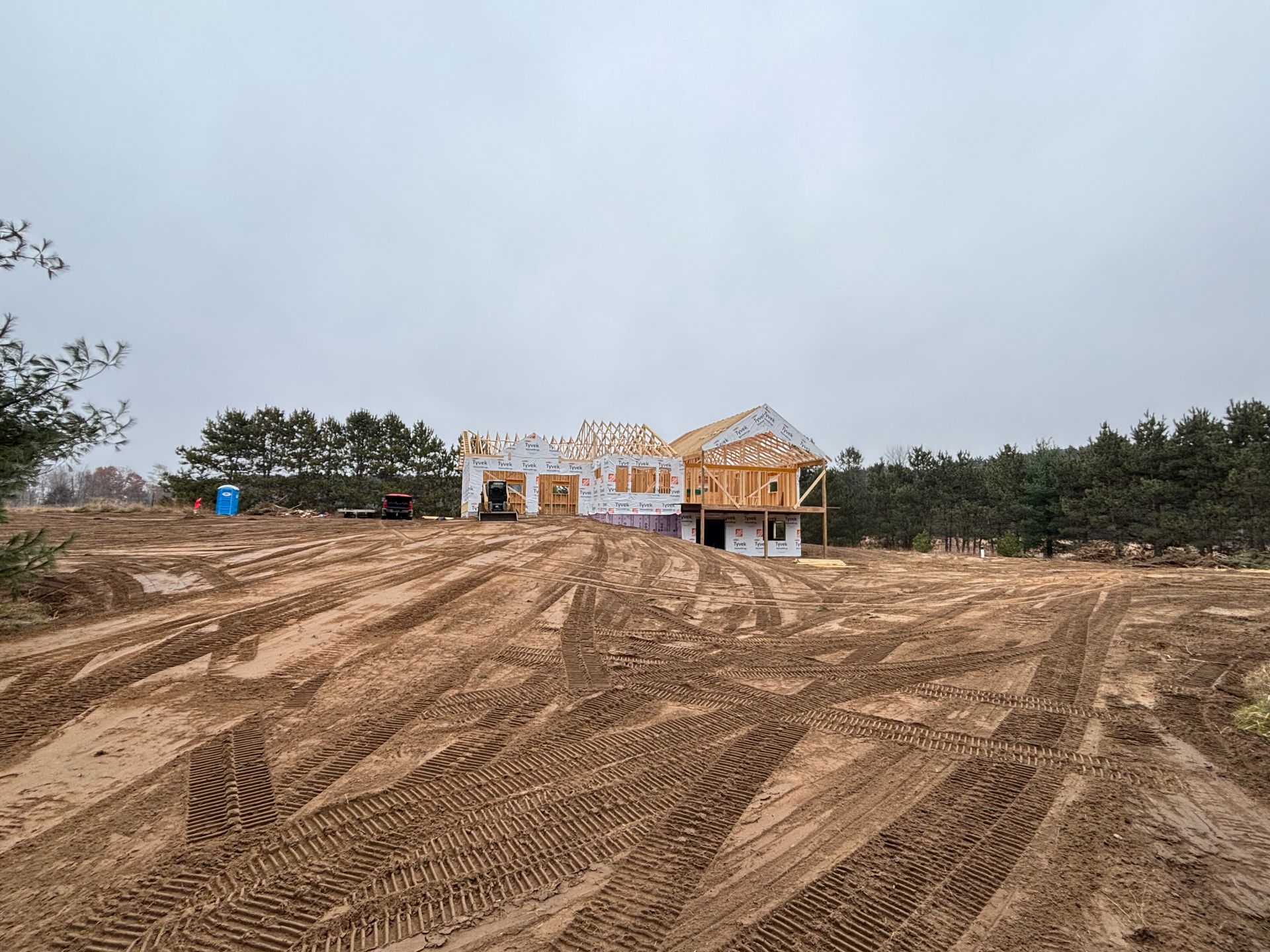 A house under construction stands in a muddy, uneven field under a cloudy sky.