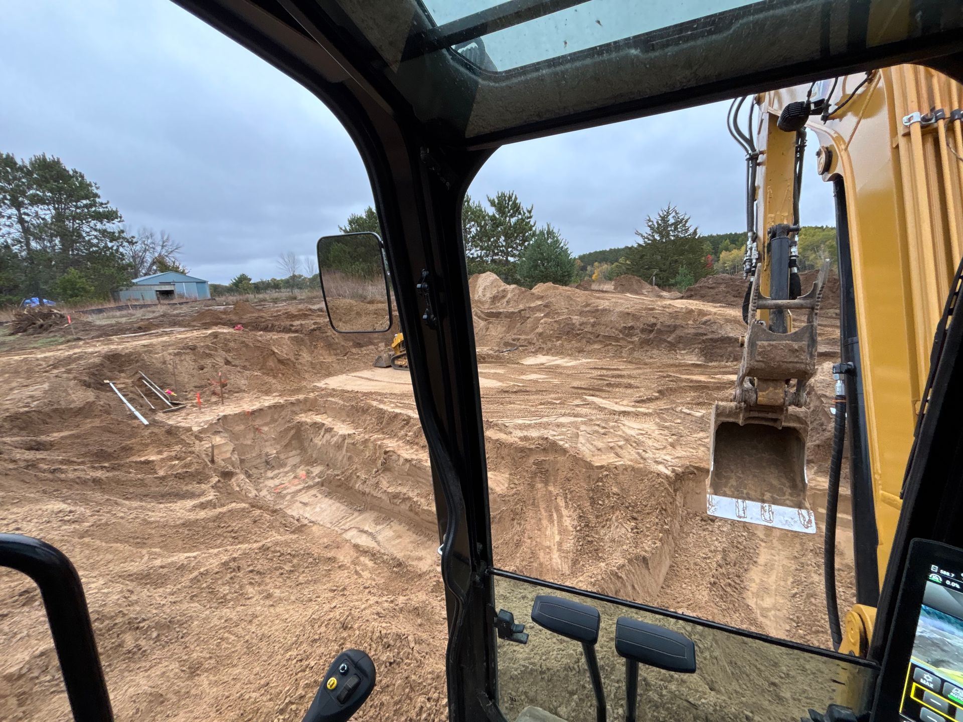 View from the cab of a yellow excavator overlooking a construction site with excavated dirt mounds on a cloudy day.