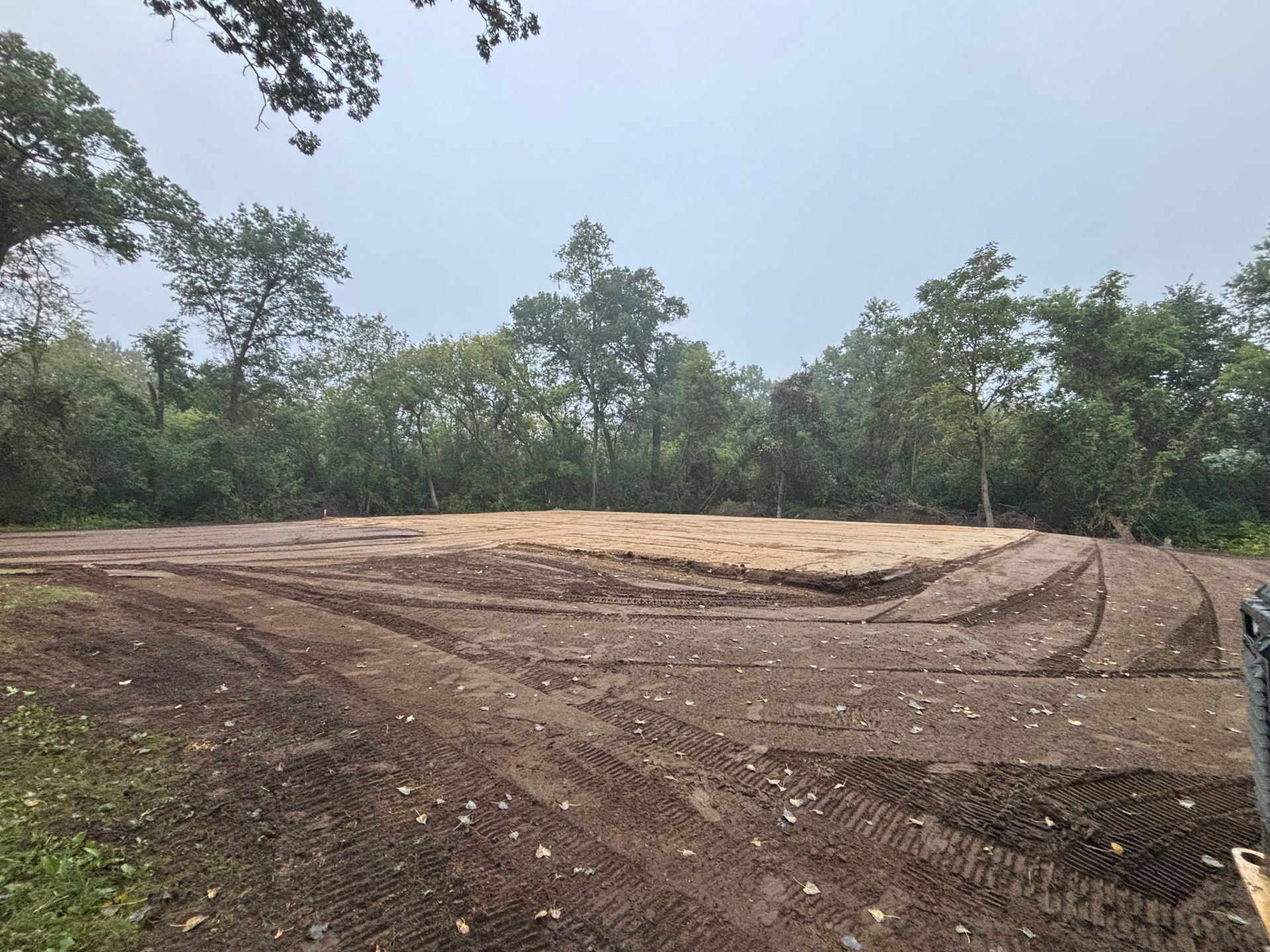 A cleared, leveled dirt construction site surrounded by a dense forest under a cloudy sky.