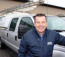 Un homme en veste bleue sourit près d'un camion de chantier surmonté d'une échelle.