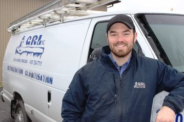 Un homme en veste bleue sourit à côté d'une camionnette blanche portant le logo « GRI Inc », un porte-échelle et un texte concernant la réfrigération.