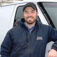 Un homme en veste et casquette bleues sourit près d'une camionnette blanche.