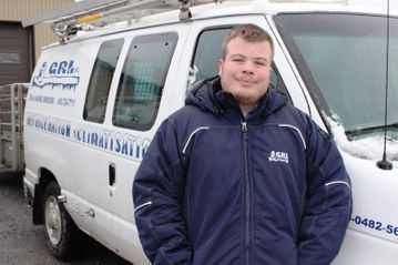 Un homme en veste bleue se tient devant une camionnette blanche arborant le logo de l'entreprise ; il neige au sol.