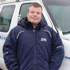Un homme en veste bleue se tient près d'un véhicule blanc.