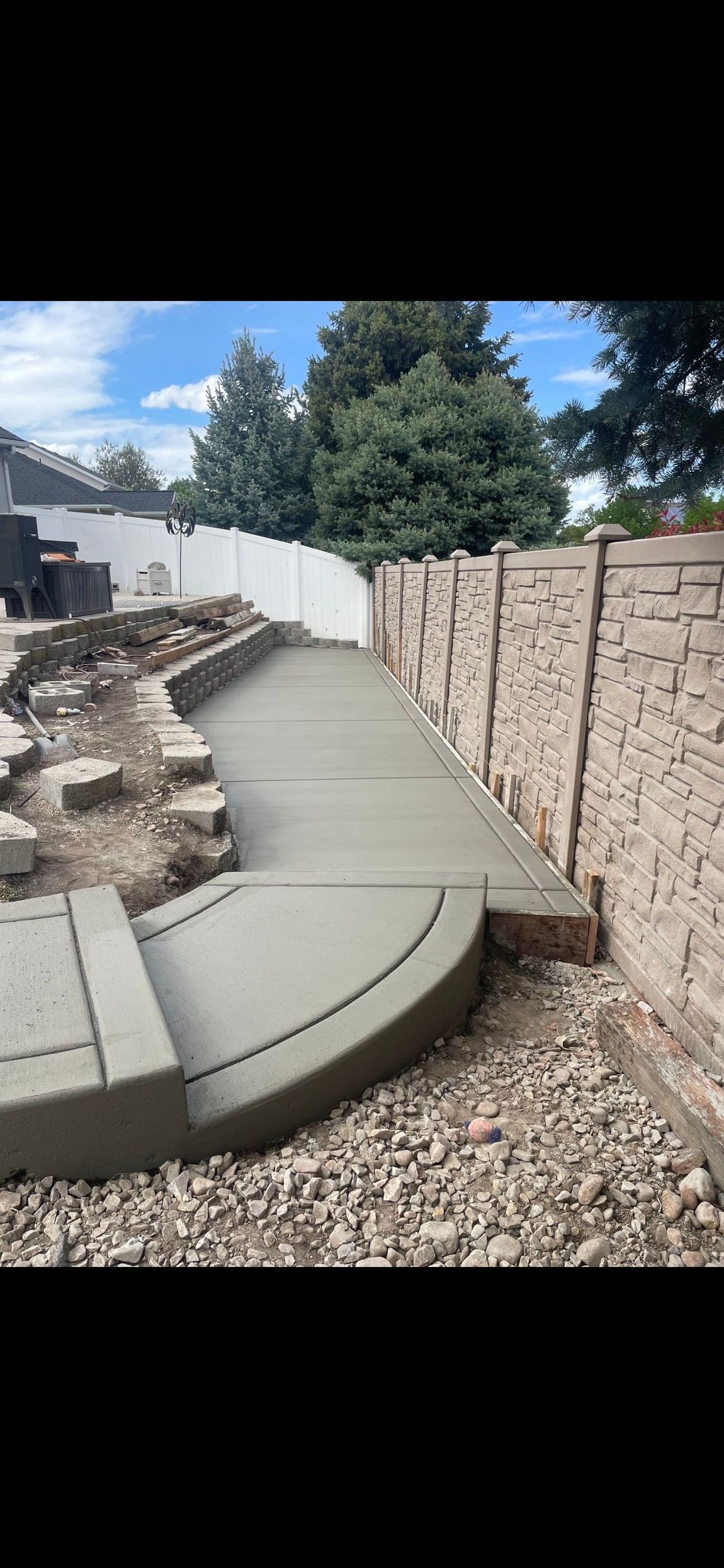 Curved concrete walkway beside a stone retaining wall and gravel yard under a blue sky
