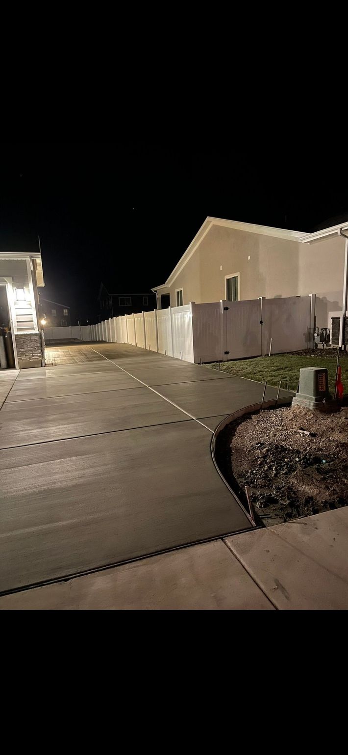 Nighttime suburban street with lit houses, a driveway, and a trash bin by the curb.
