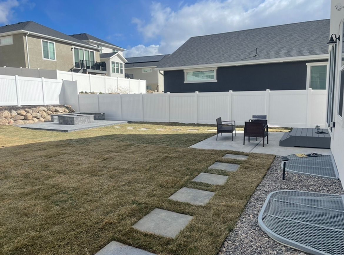 Backyard patio with stone path, lawn, white fence, and outdoor chairs under a blue sky