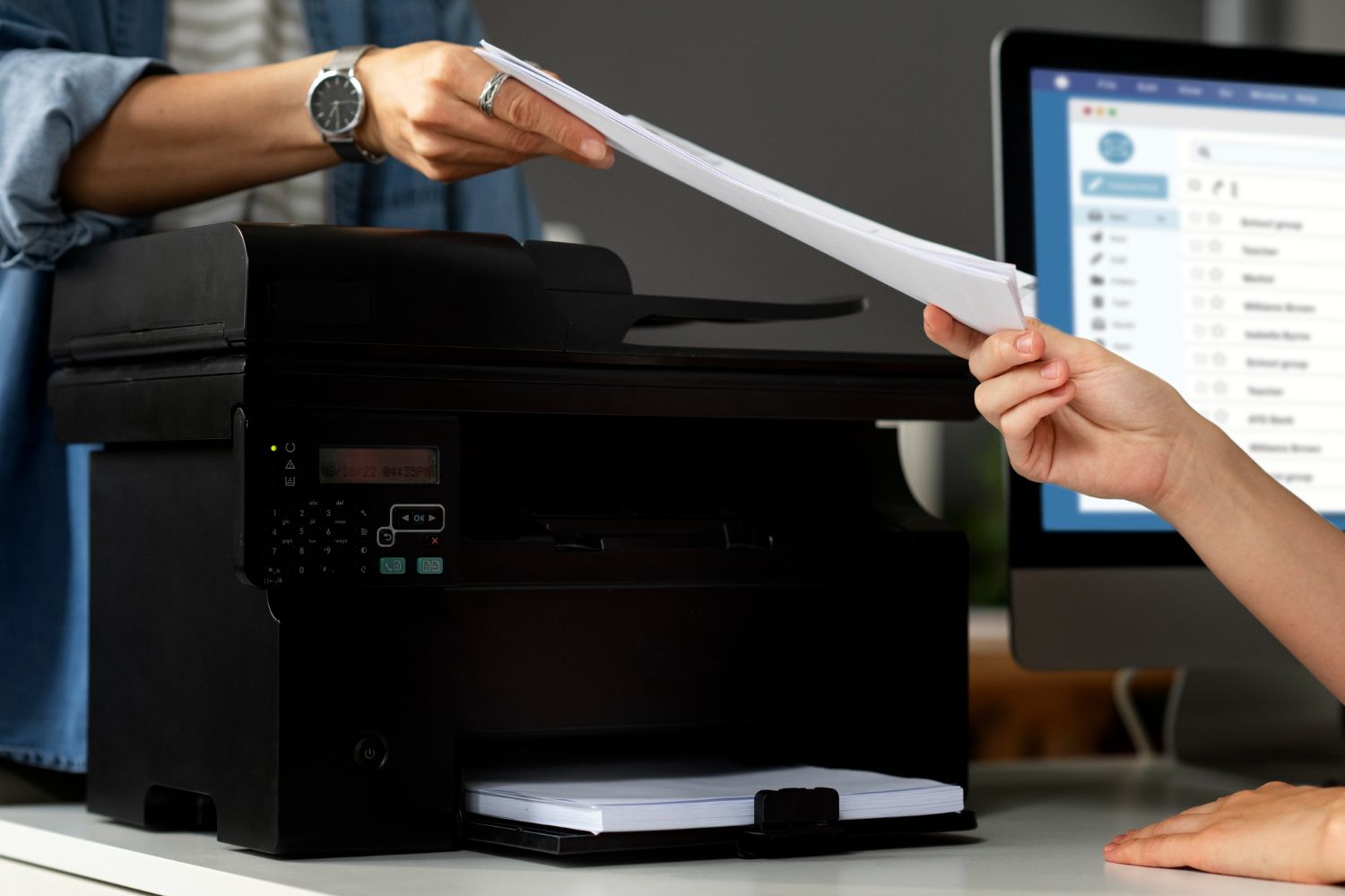 Dos personas se pasan una pila de papel blanco por encima de una impresora de oficina negra, cerca de un monitor de ordenador.