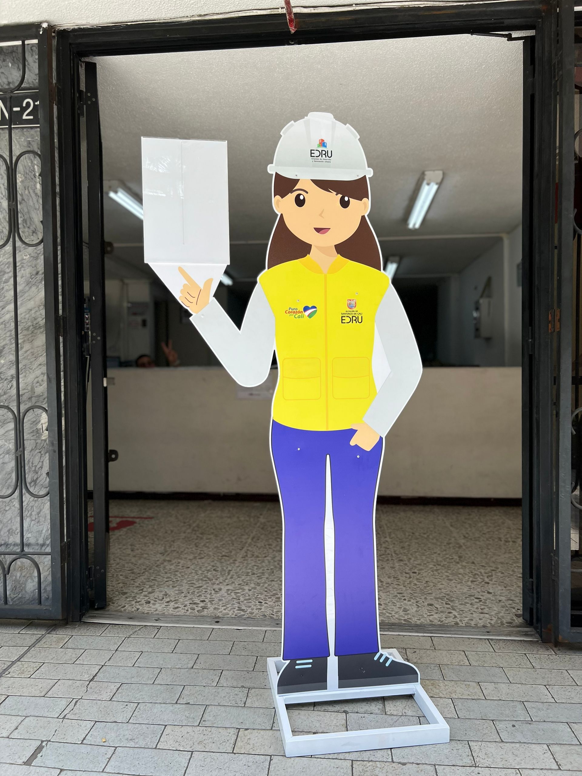 Dibujo animado de un obrero de la construcción con chaleco amarillo y casco blanco, de pie en una puerta, sosteniendo unos papeles.