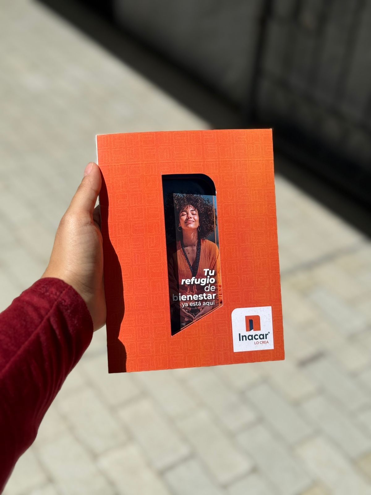 Mano sosteniendo un folleto naranja con una pequeña foto en la portada, sobre un fondo de pavimento borroso.