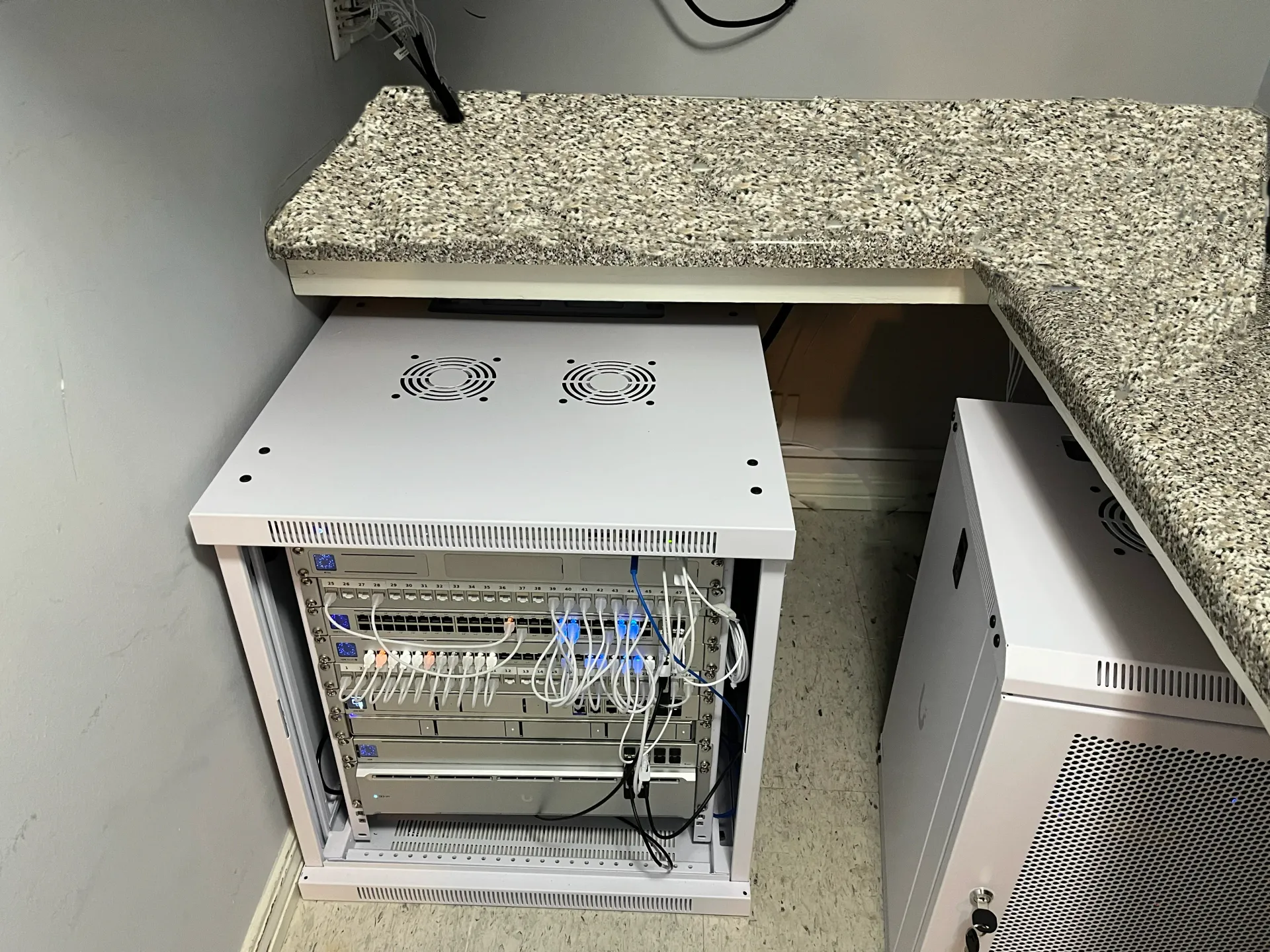 unifi switches, routers, nas & patch panels mounted to floor standing white rack. devices are cabled, powered on & showing activity leds with another rack on the side