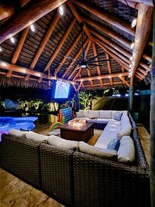 Outdoor lounge with thatched roof, sectional sofa, fire pit, and pool at night.